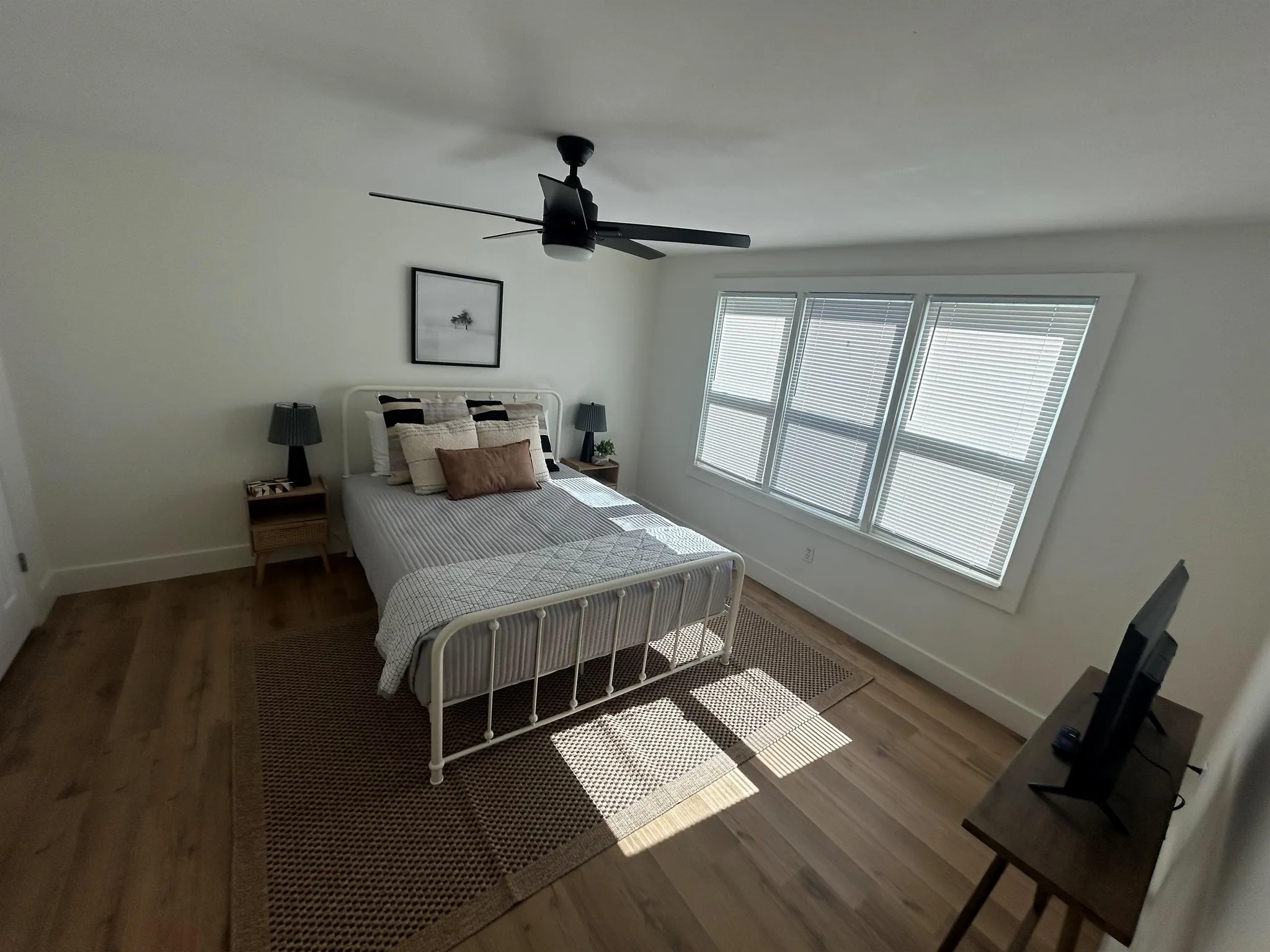 A bedroom with a white metal bed, a black ceiling fan, and large windows.