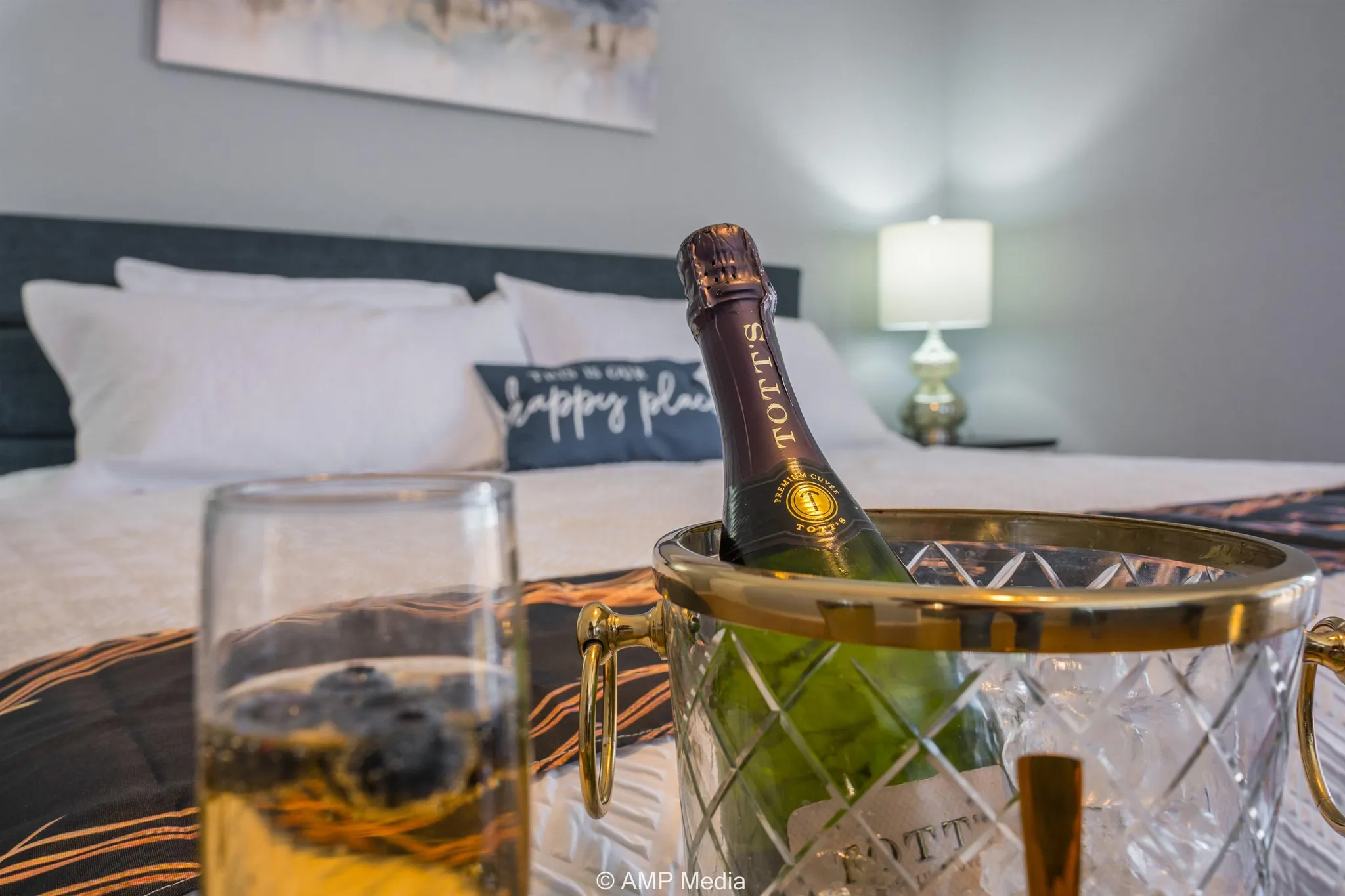 A bottle of champagne chills in an ice bucket next to a glass of champagne on a bed.
