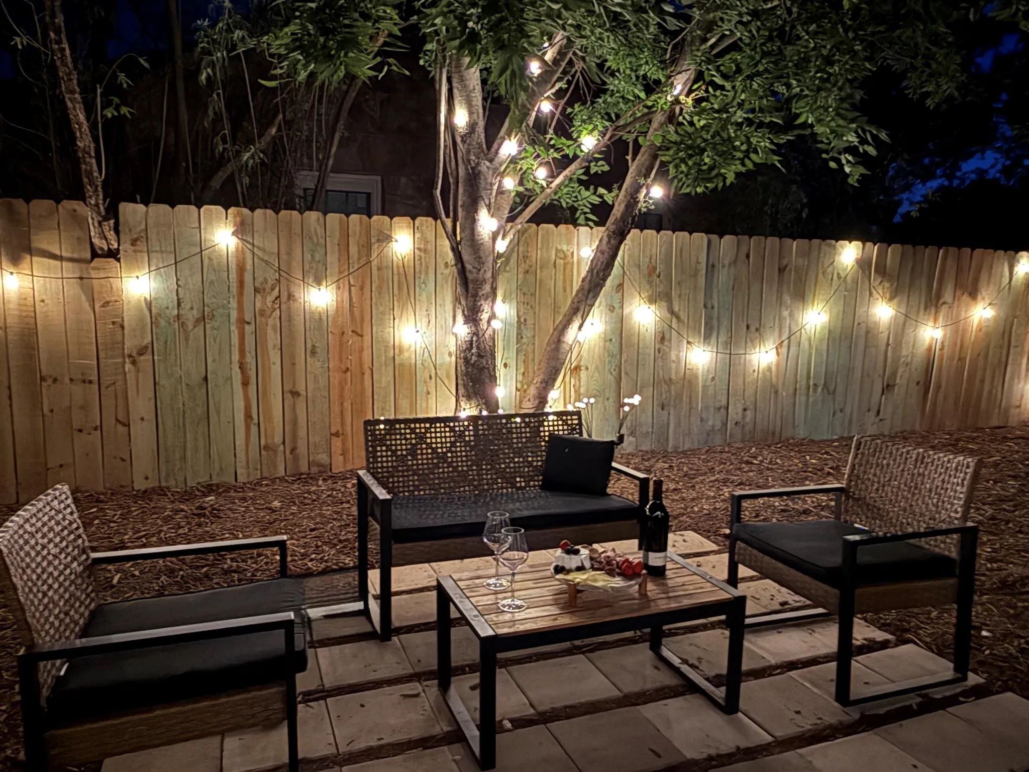 A nighttime backyard patio scene with string lights, seating, and a small table with wine and snacks