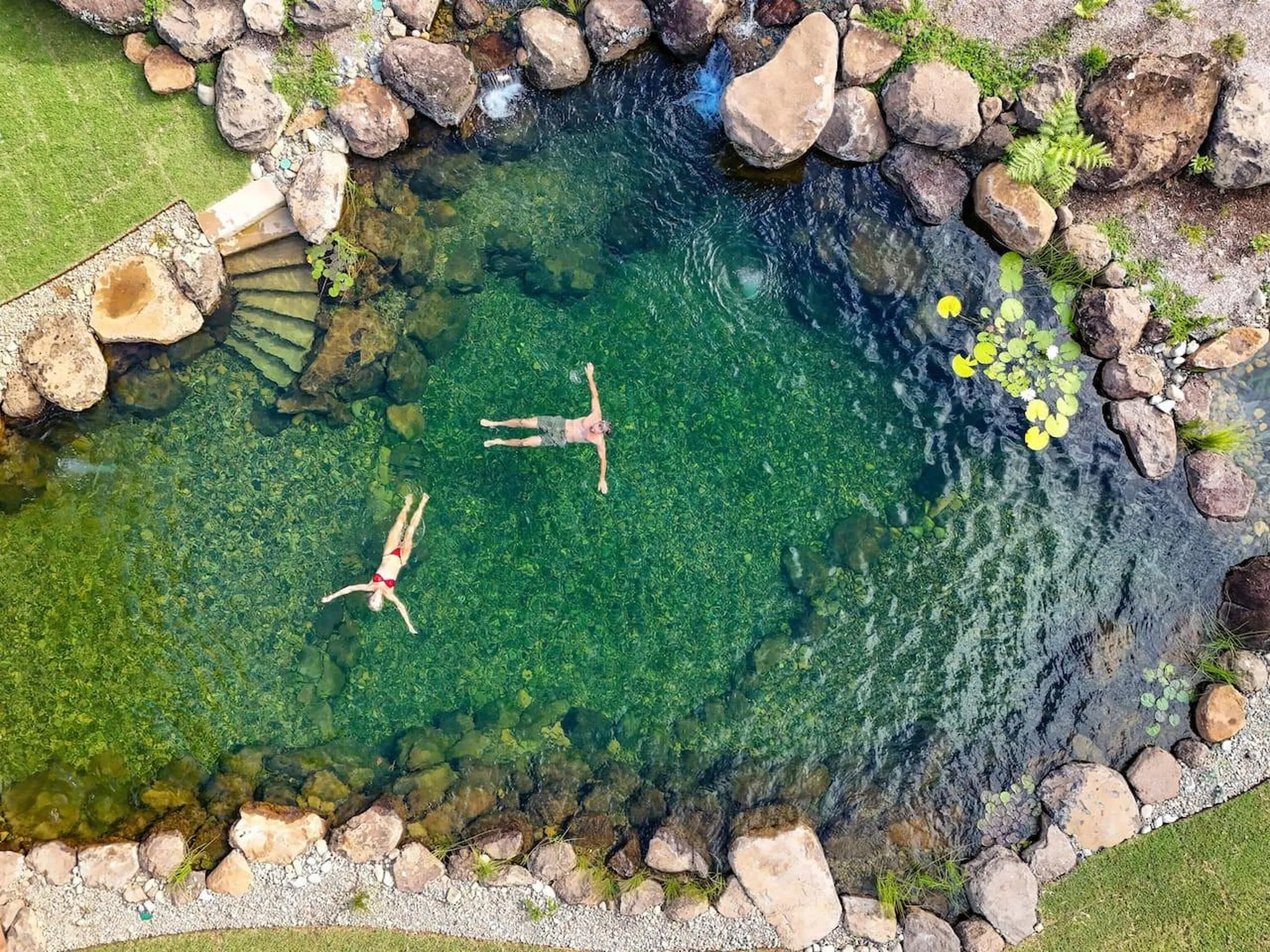 Two people float in a clear green pond surrounded by rocks.