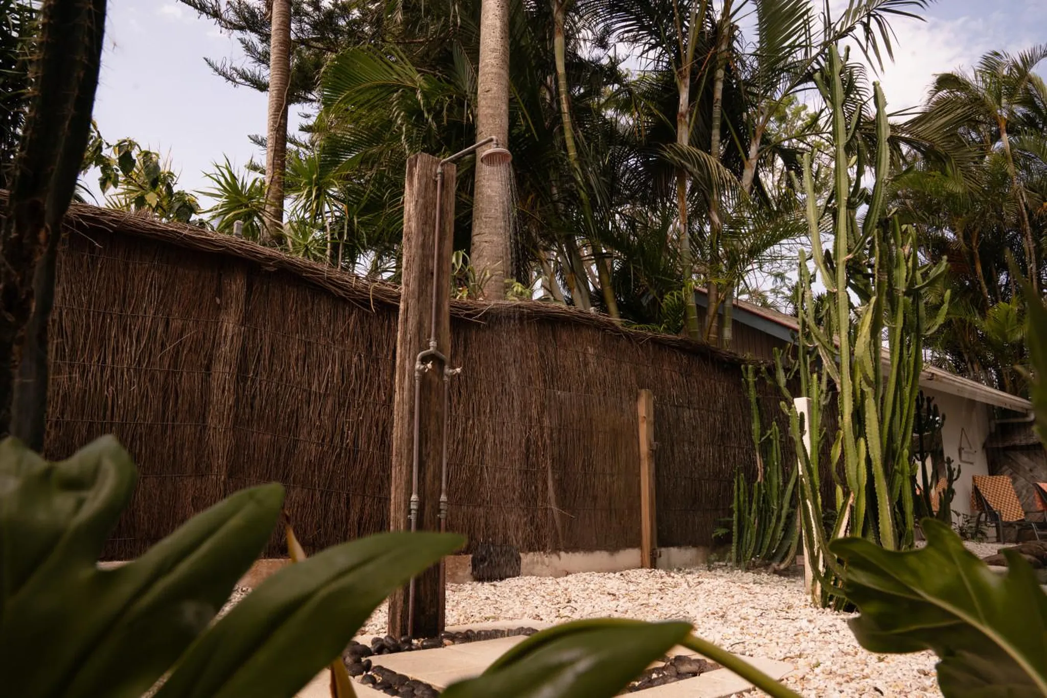 Outdoor shower with wooden post, against a thatched wall and lush tropical foliage.
