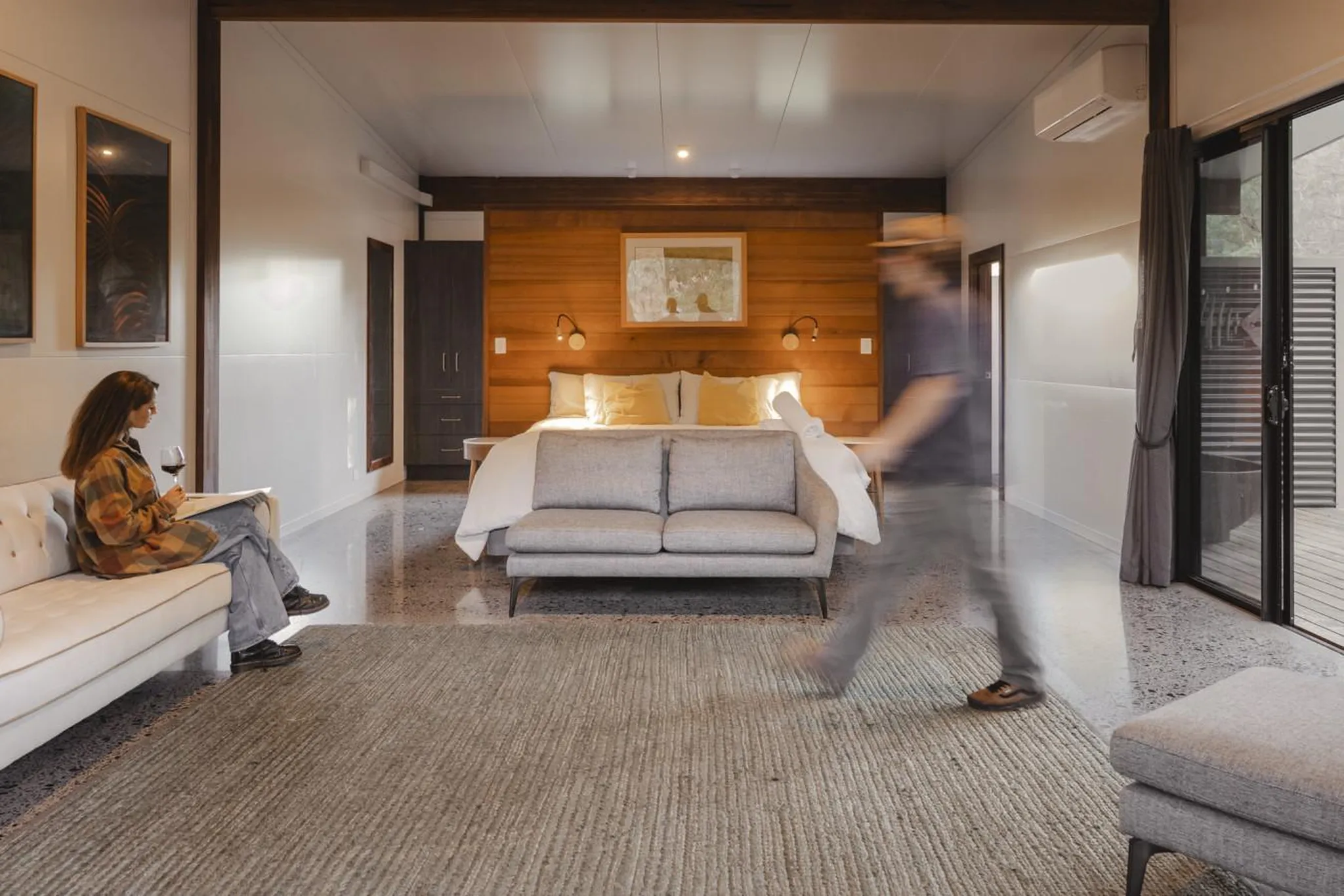 A person walks past a bed with a grey couch in front of it in a room with wooden walls.