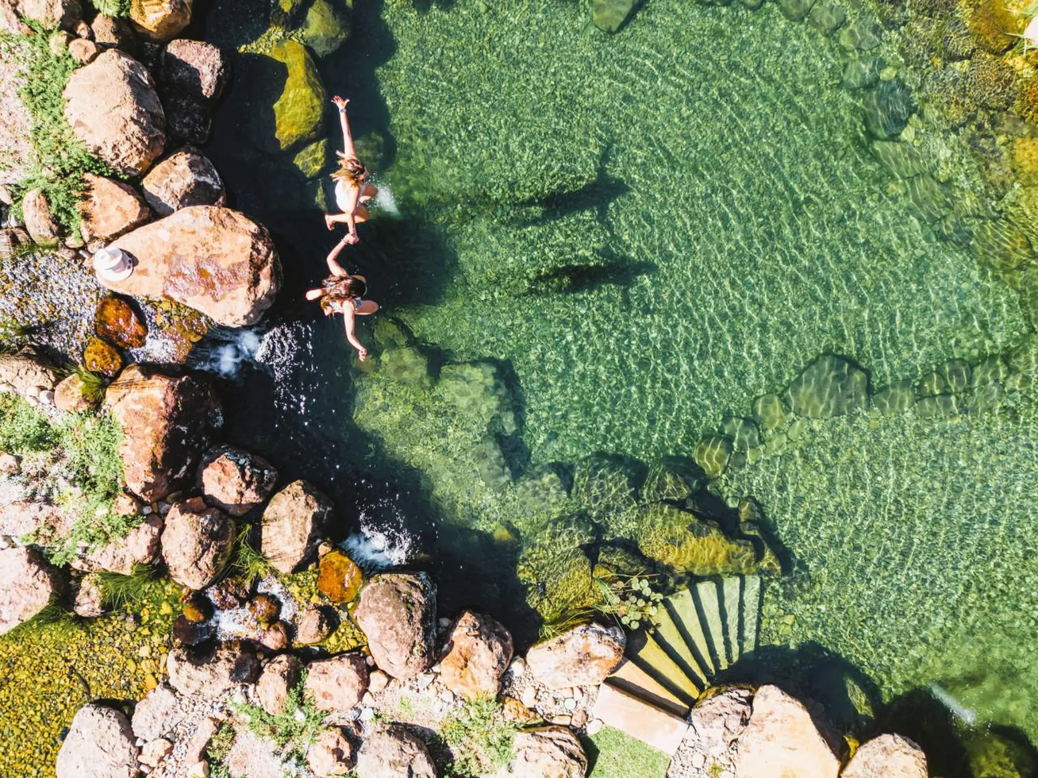 Aerial view of two women jumping into a clear, shallow pool with rocky edges.