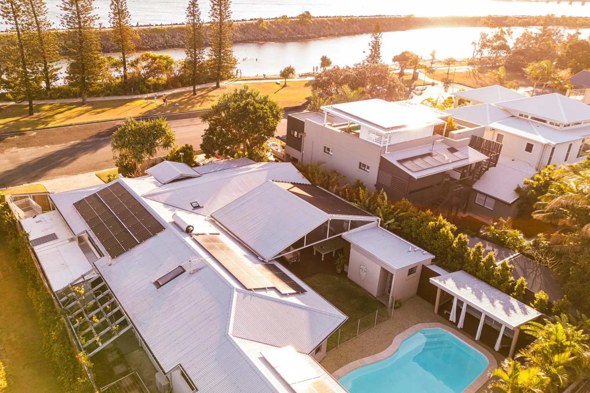 Aerial view of houses with solar panels and a swimming pool near a body of water at sunset.