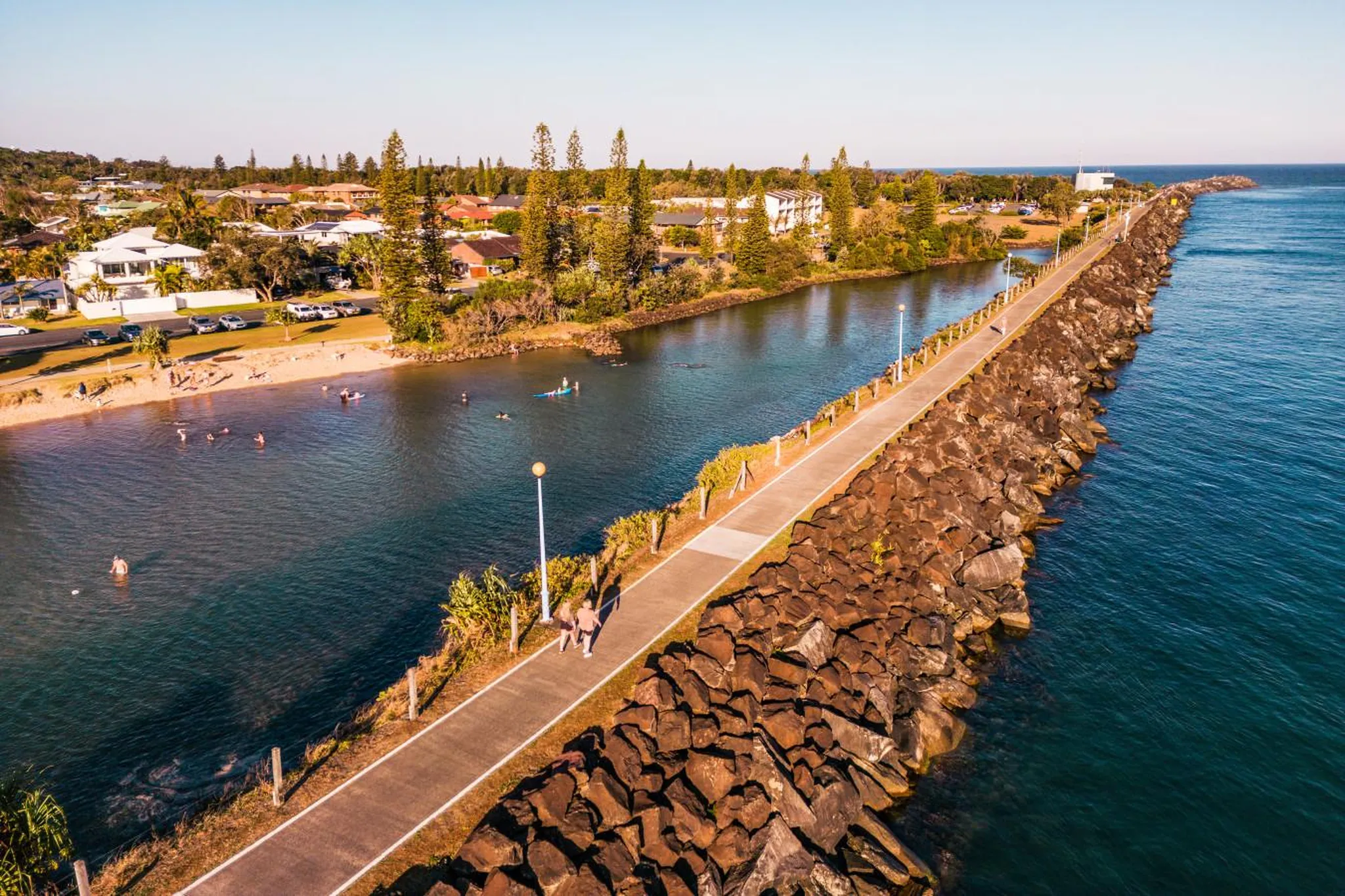 An aerial view of a paved walkway along a rocky breakwater with people walking and swimming in the w