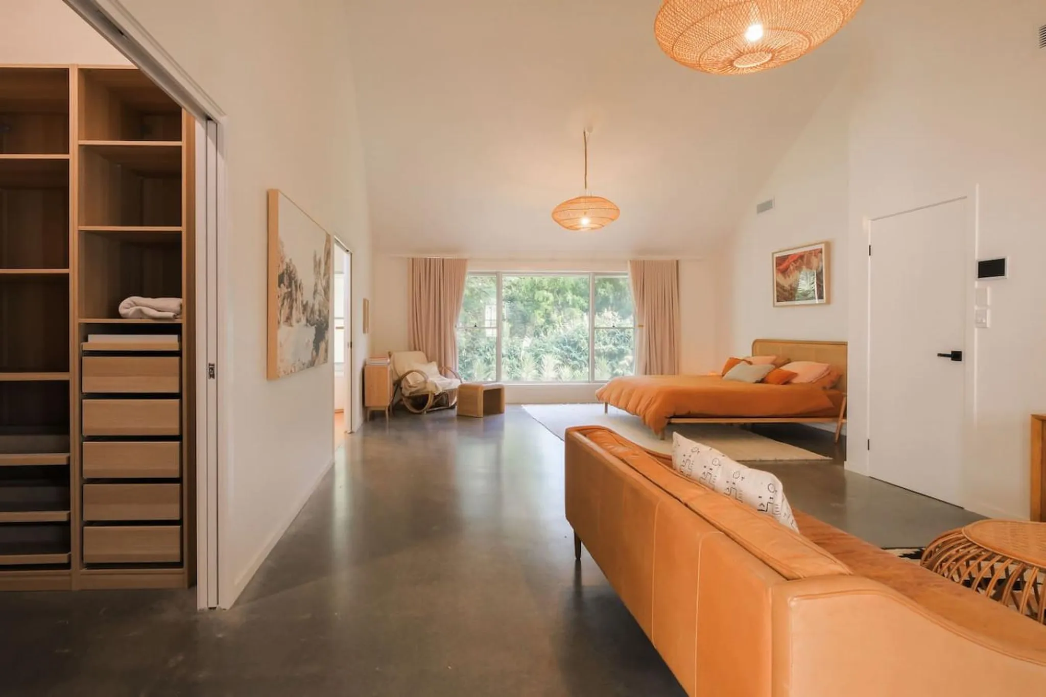 Bedroom with orange bed, tan couch, and large window with lush greenery outside.