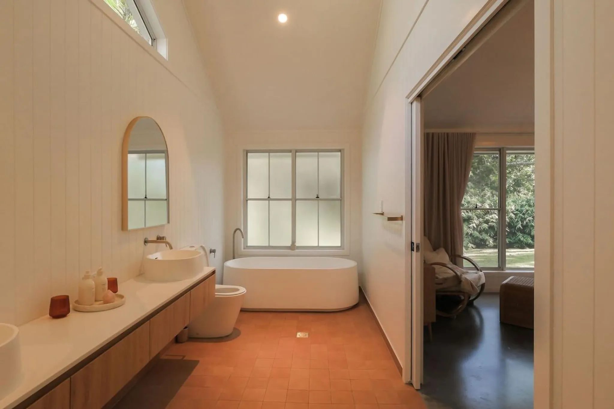 A bright, minimalist bathroom with a freestanding tub, vanity, and toilet, opening to a bedroom with