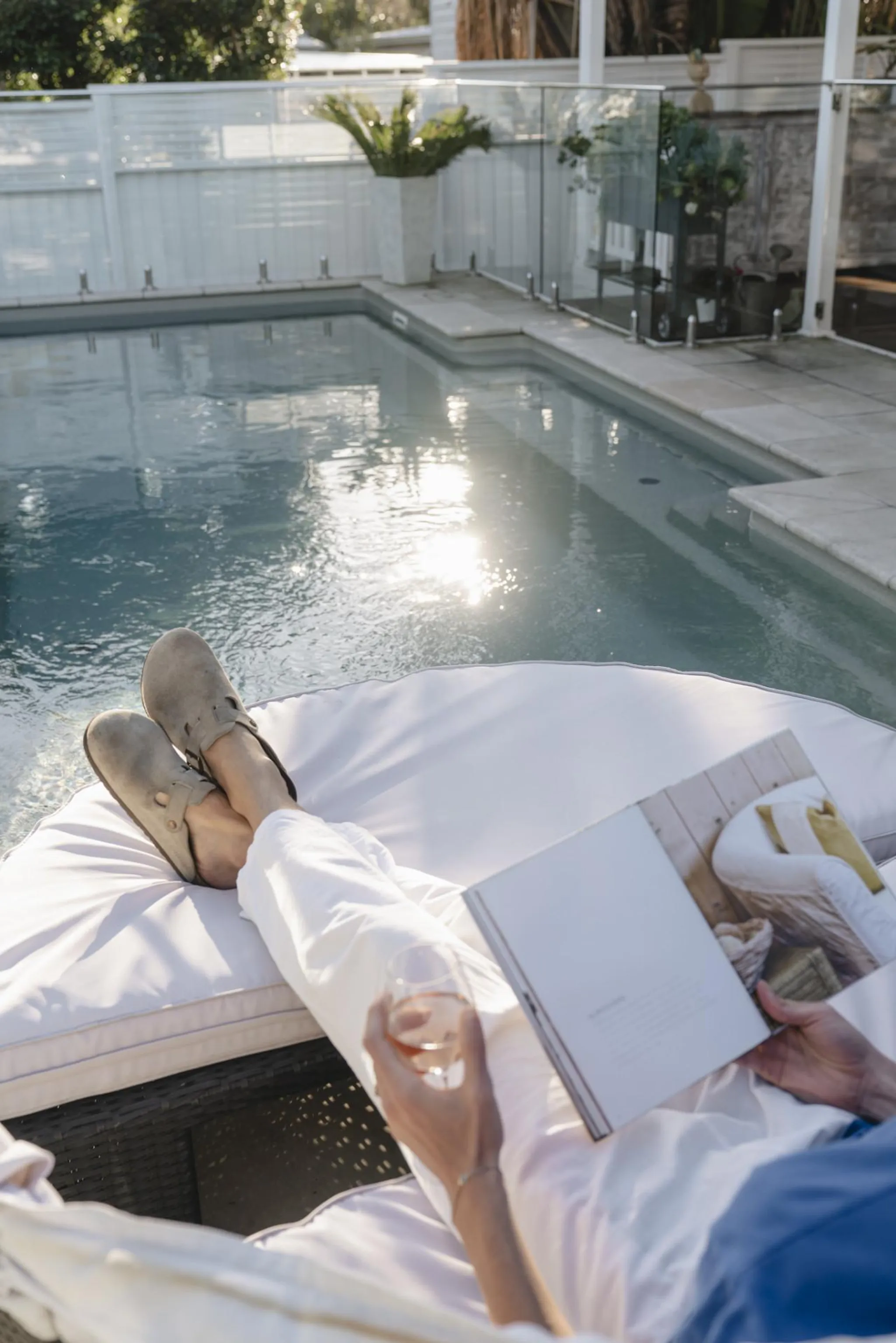 A person relaxing by a pool reads a book and holds a drink.