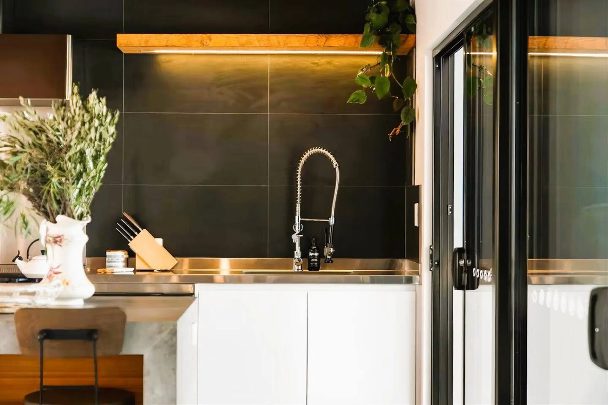 A modern kitchen sink with black tiles, a stainless steel countertop, and a wooden shelf.