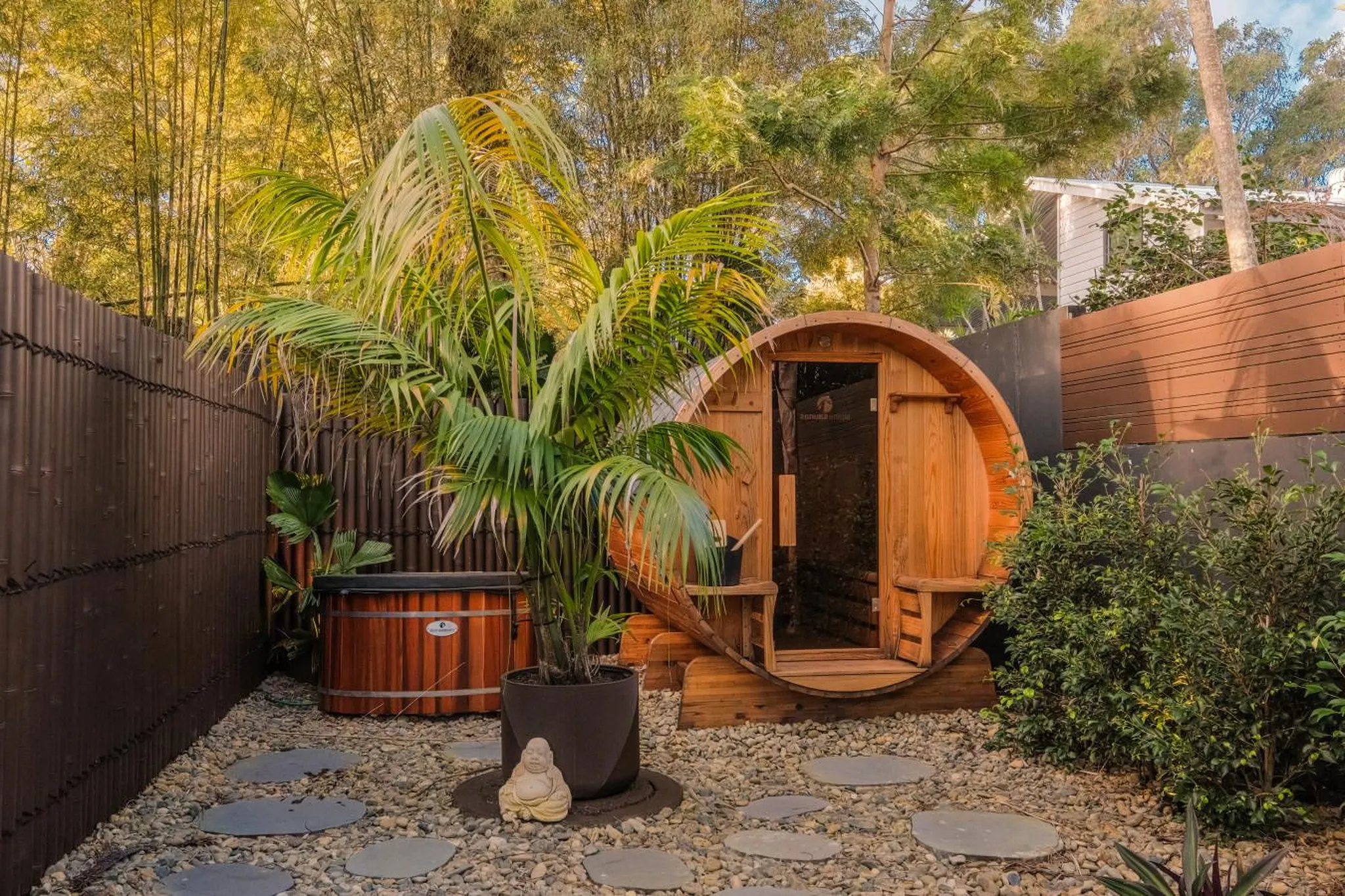 Wooden barrel sauna and hot tub in a tranquil backyard with bamboo and lush greenery.