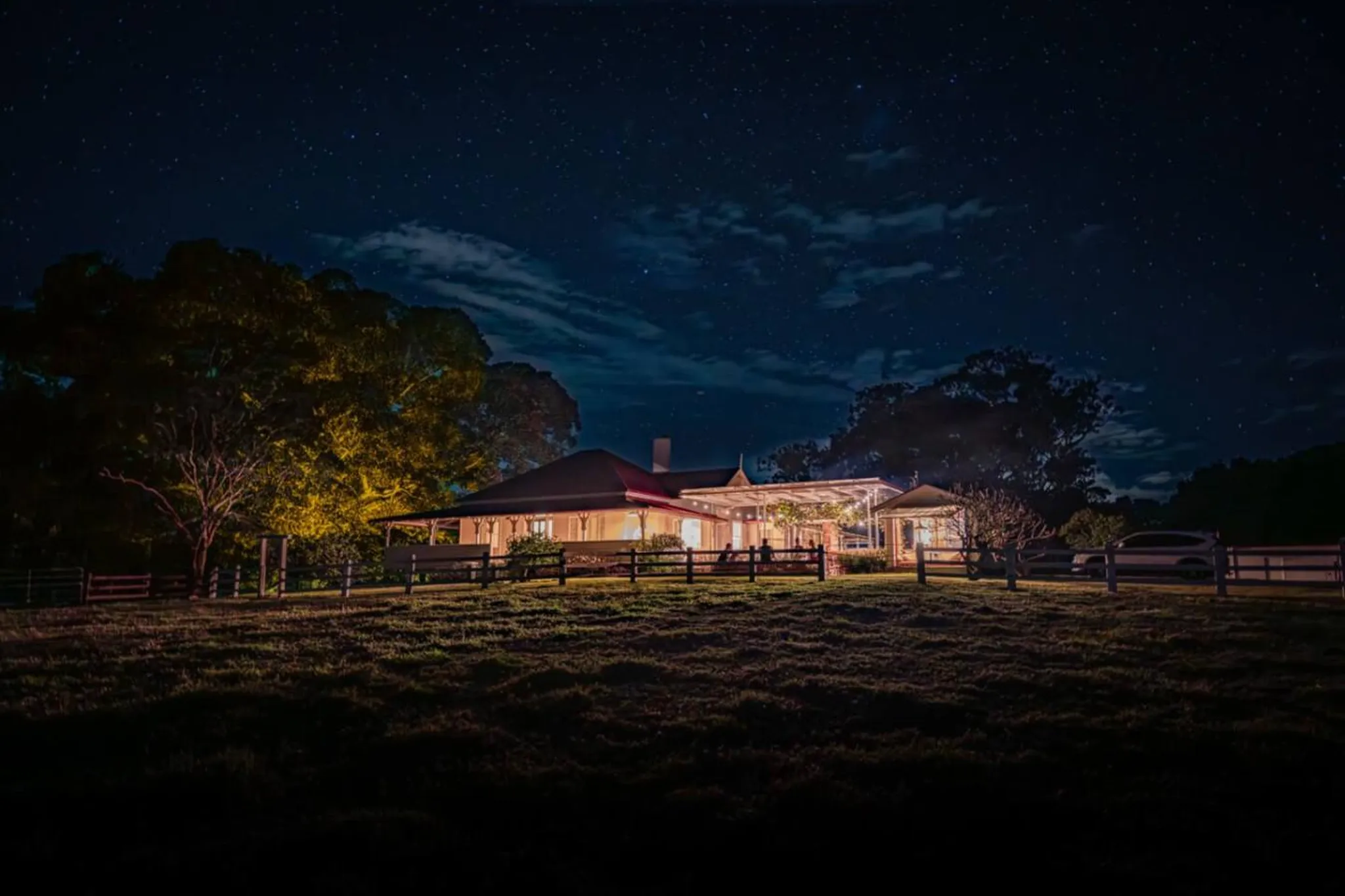 A large, lit-up house at night under a starry sky with a fence in the foreground.
