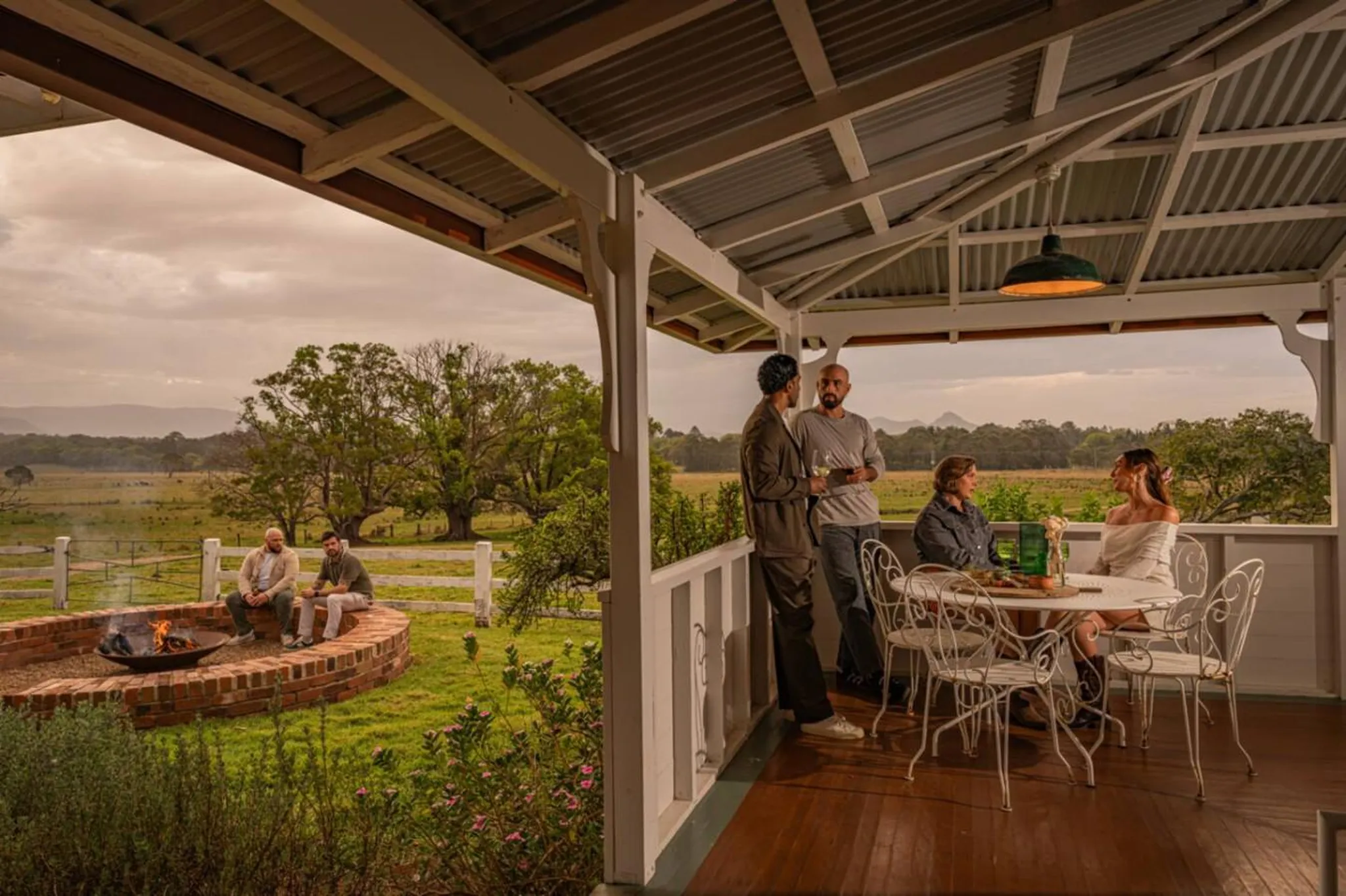 People socialize on a porch overlooking a rural landscape with a fire pit.