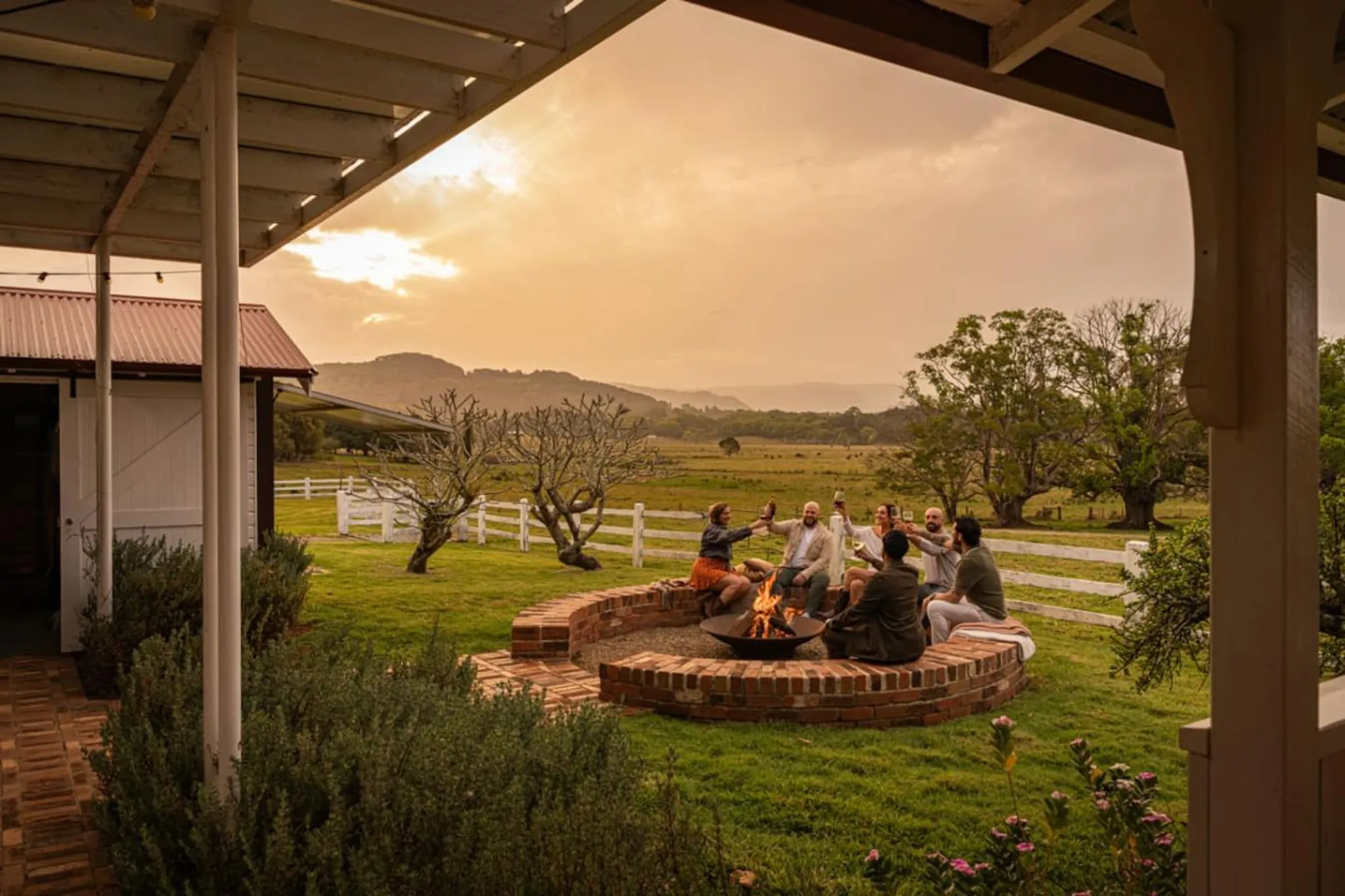 Friends gather around a fire pit at sunset, raising glasses in a toast.