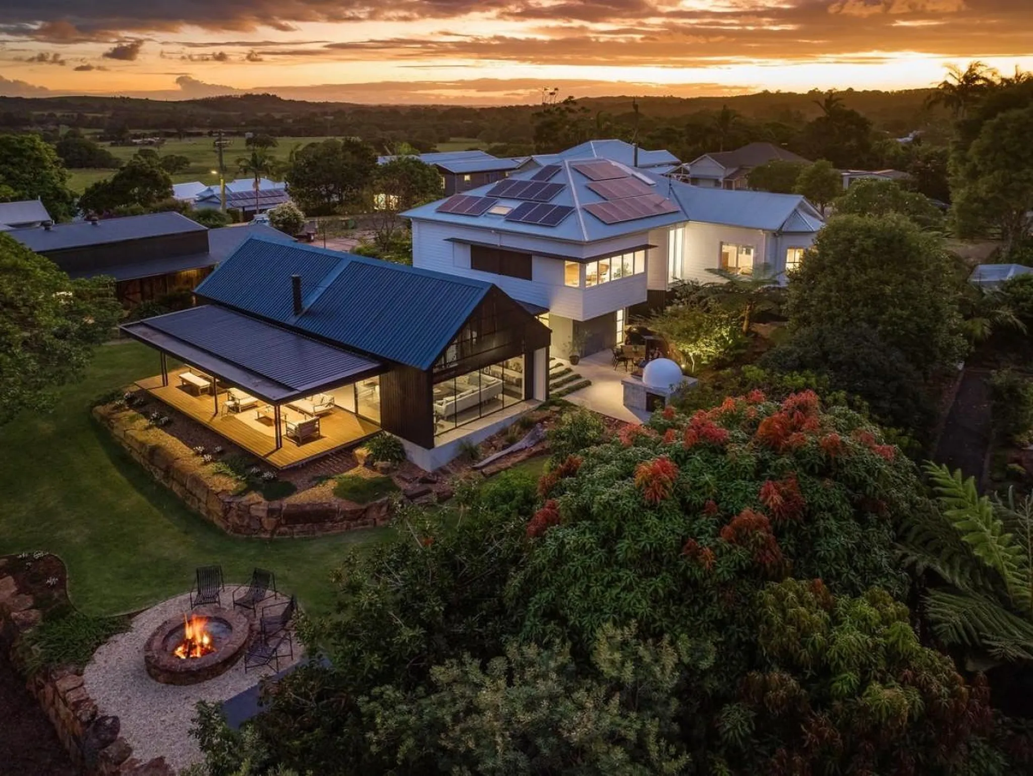 Aerial view of a modern house with a dark roof and large windows, surrounded by lush greenery and a 