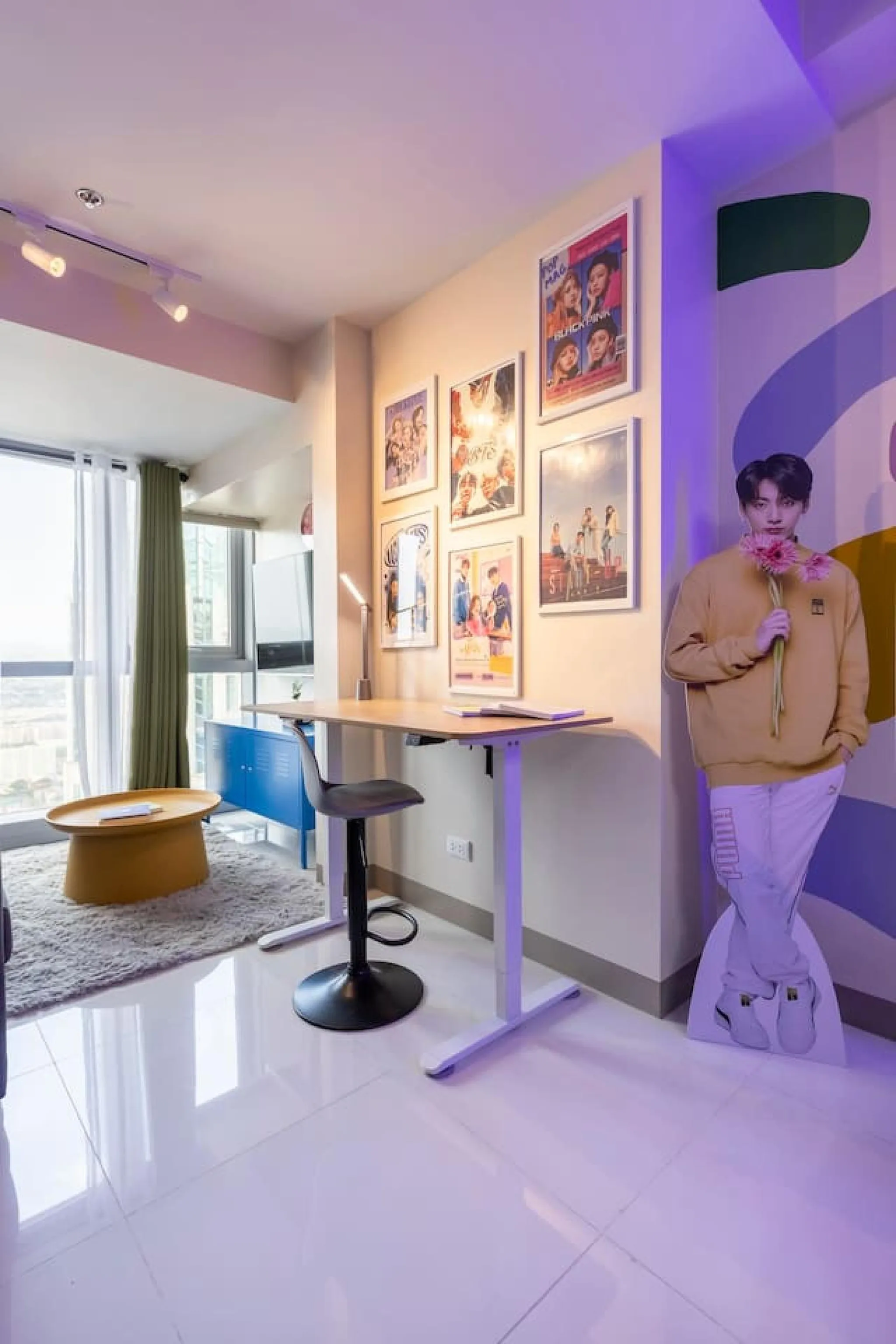 A standing desk and stool are in front of a wall adorned with framed posters.