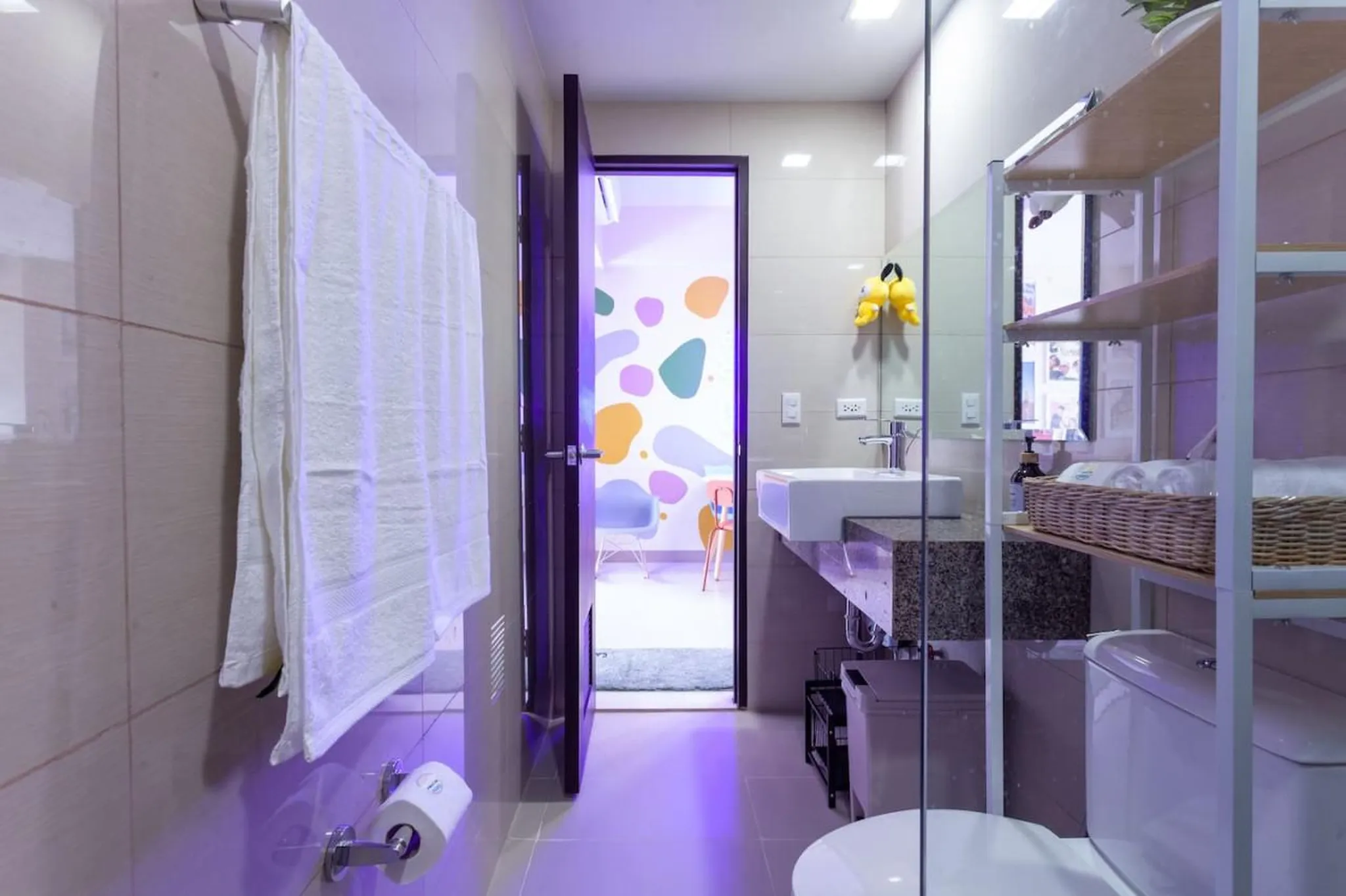 A modern bathroom with purple lighting, featuring a sink, toilet, and shelves with towels.