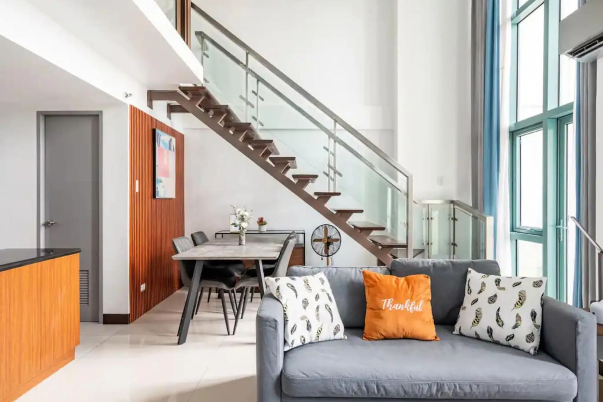 A modern living room with a gray sofa, a dining table and chairs, and a staircase with glass railing