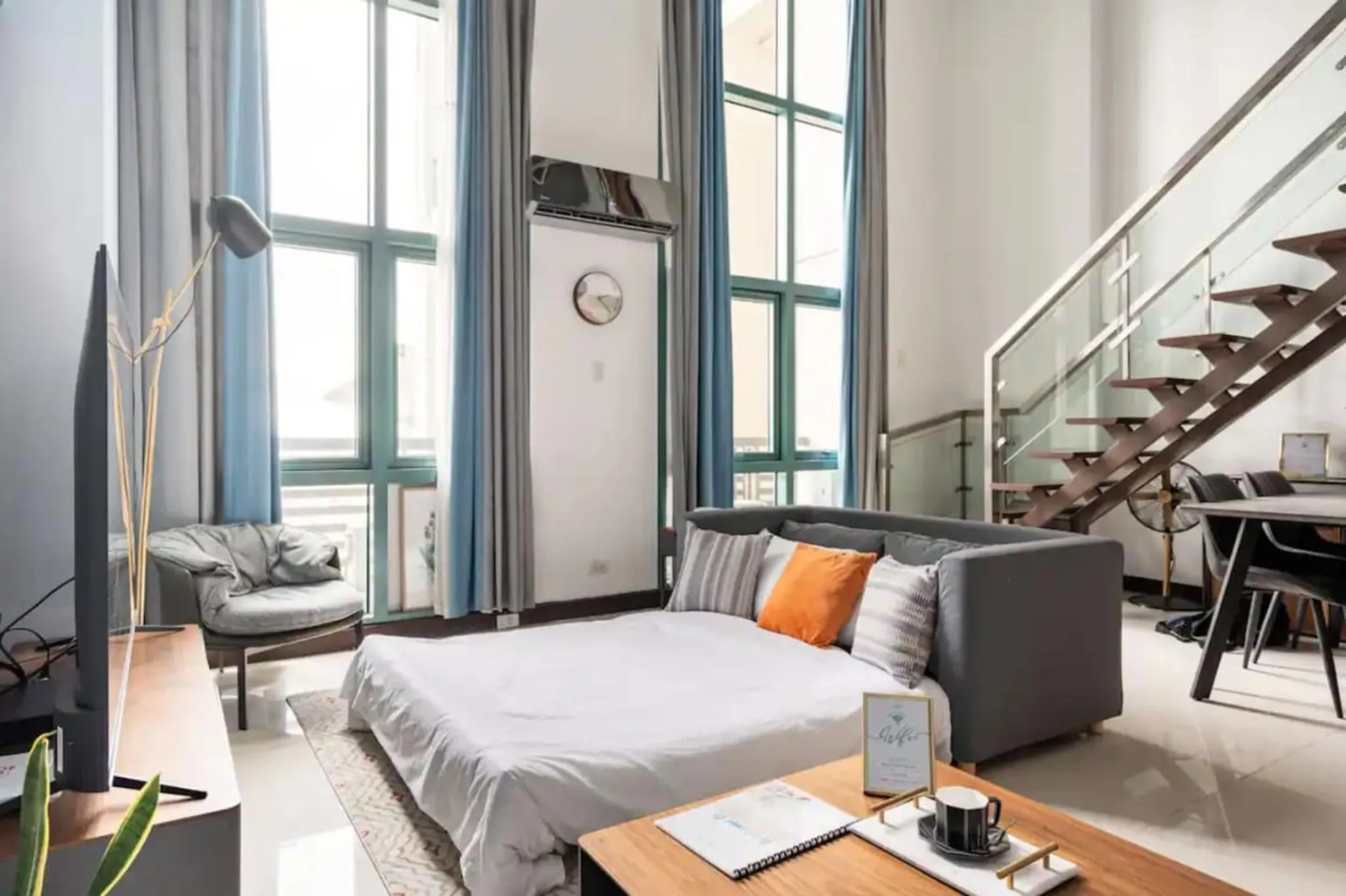 A loft bedroom with a grey sofa-bed, modern armchair, tall windows, and a wooden staircase.