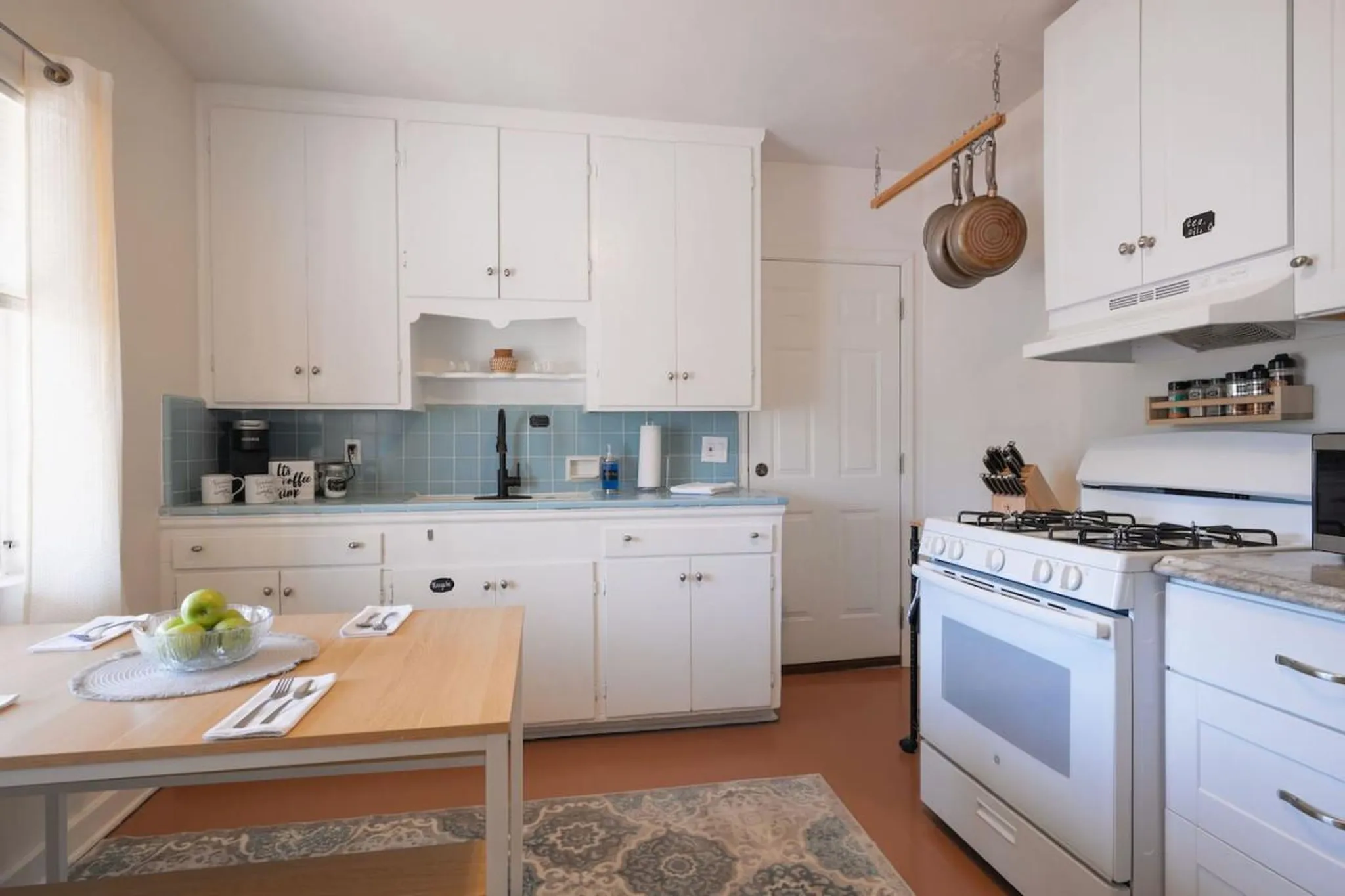 A kitchen with white cabinets, a blue tile backsplash, and a wooden table with green apples.