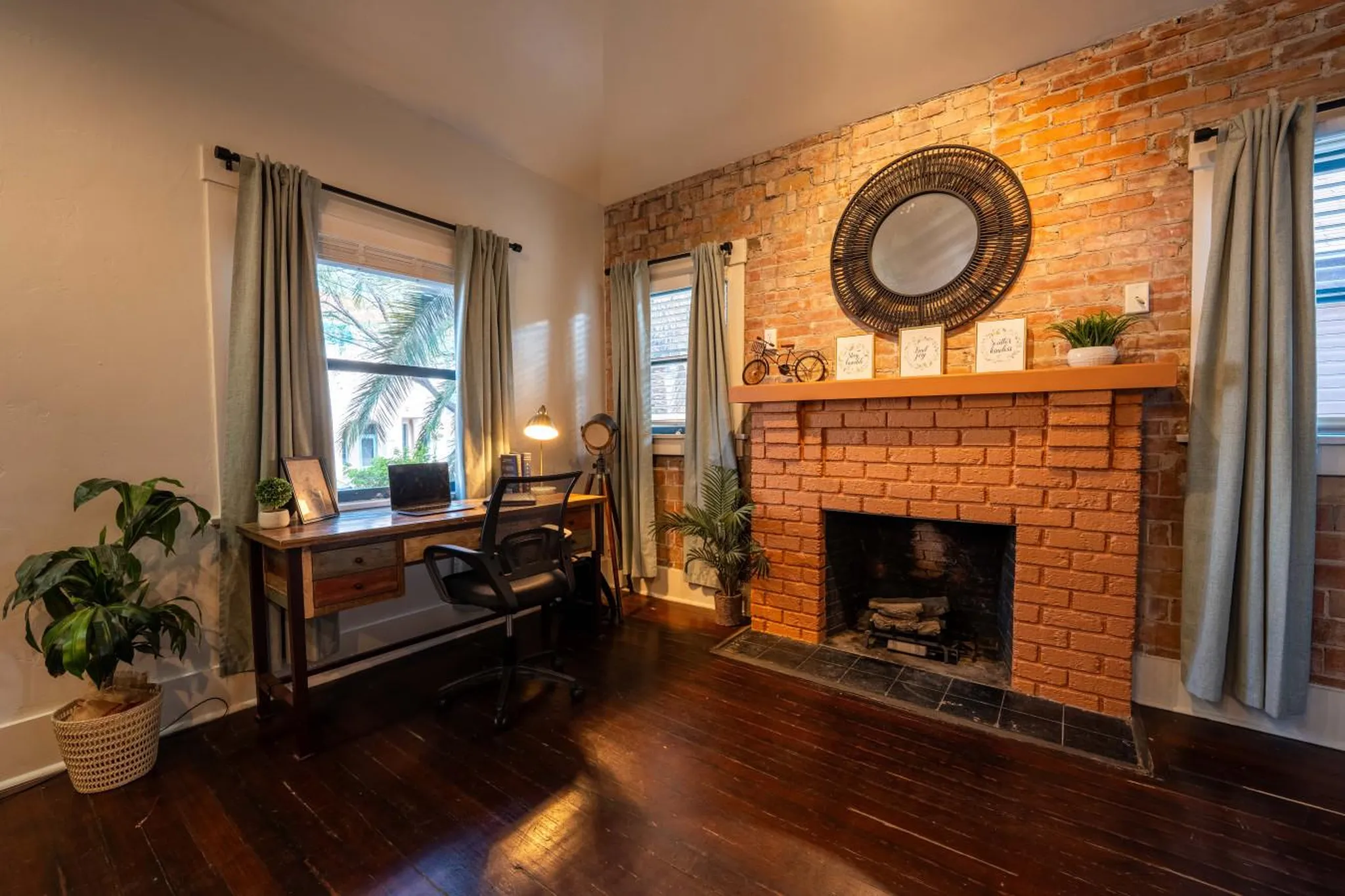 A rustic brick fireplace anchors a home office with a desk, chair, and plant.