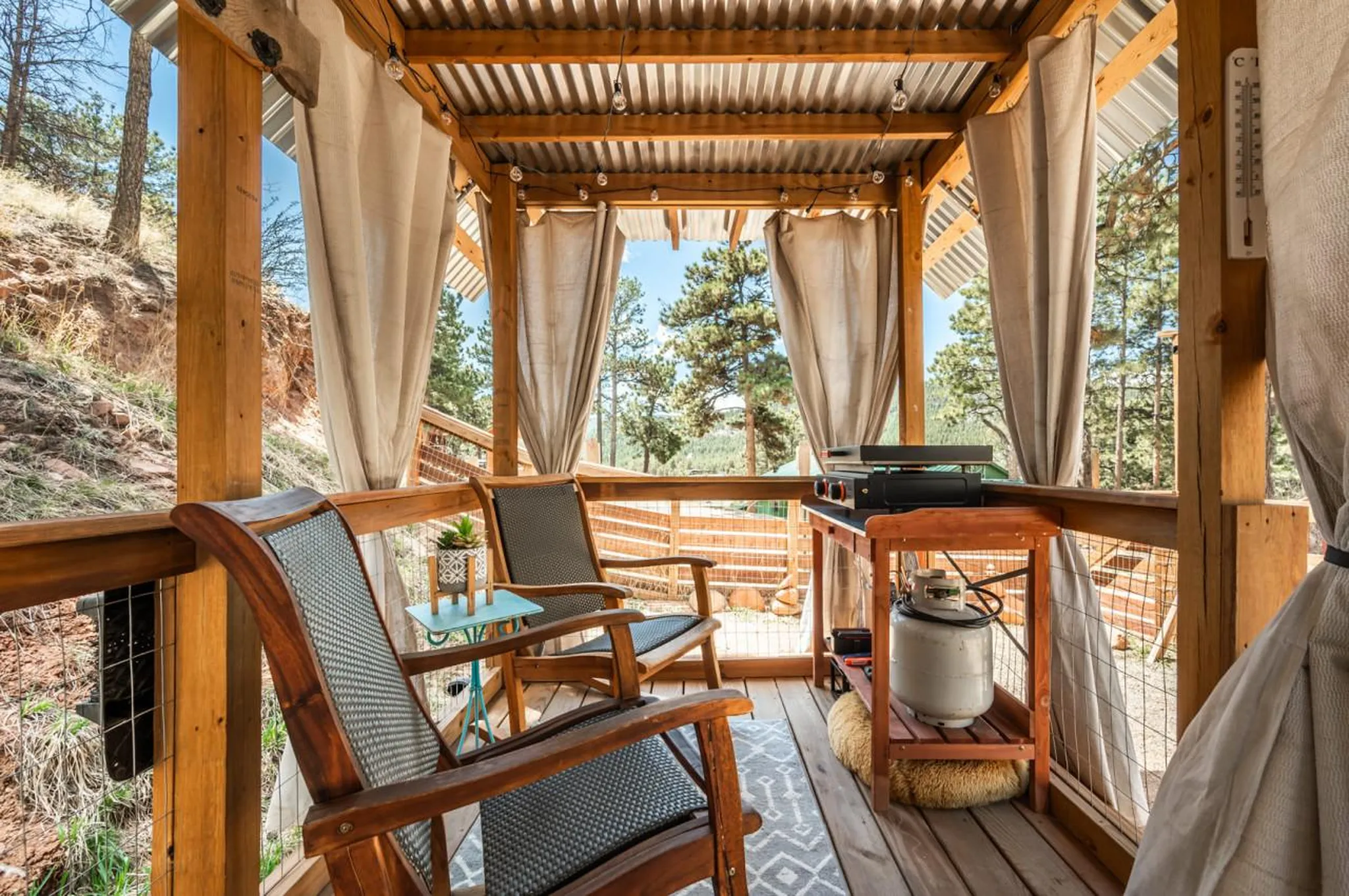 Outdoor wooden porch with chairs, a grill, and propane tank, overlooking trees.