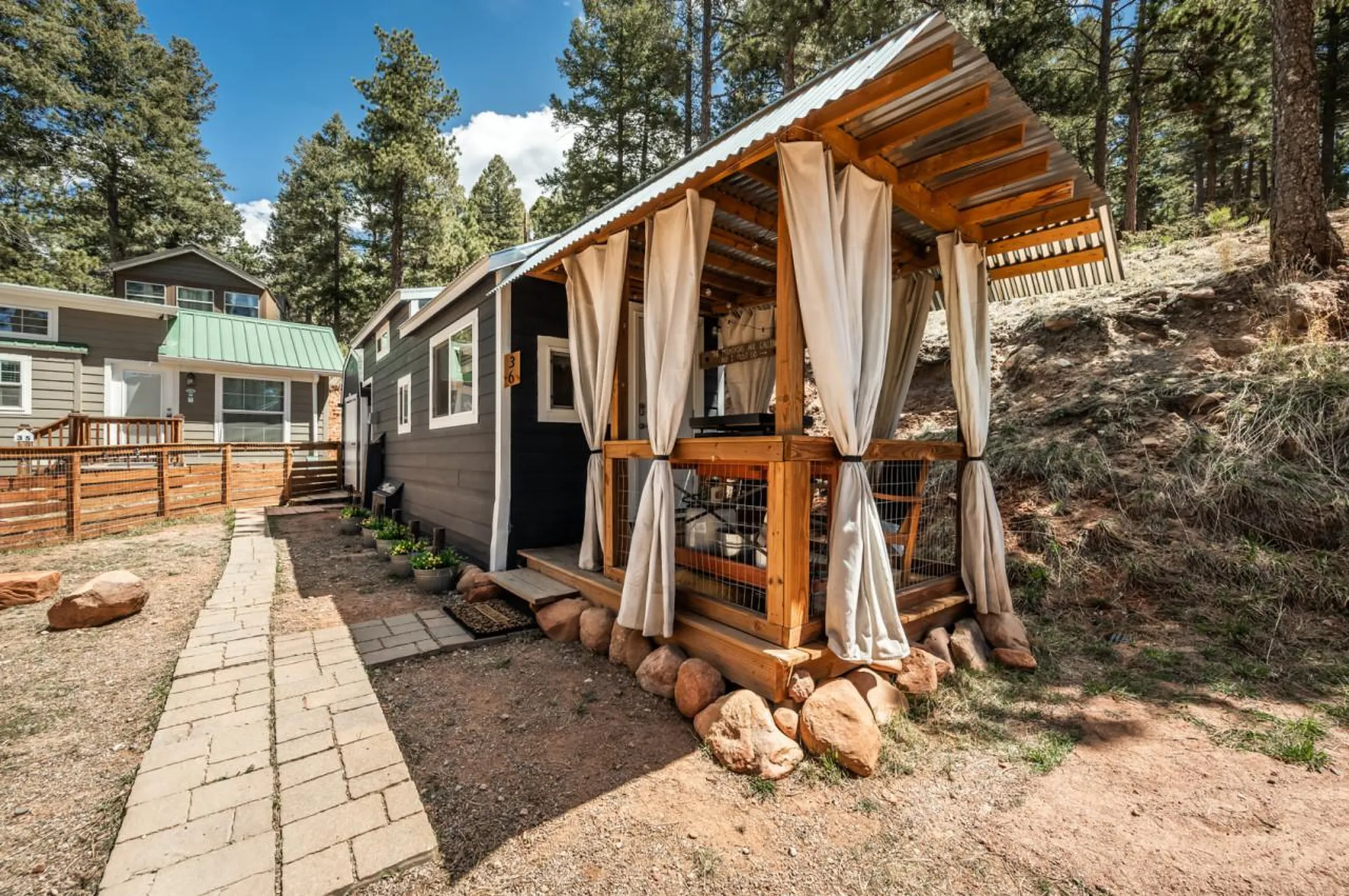A tiny home with an outdoor covered patio, a stone walkway, and pine trees.