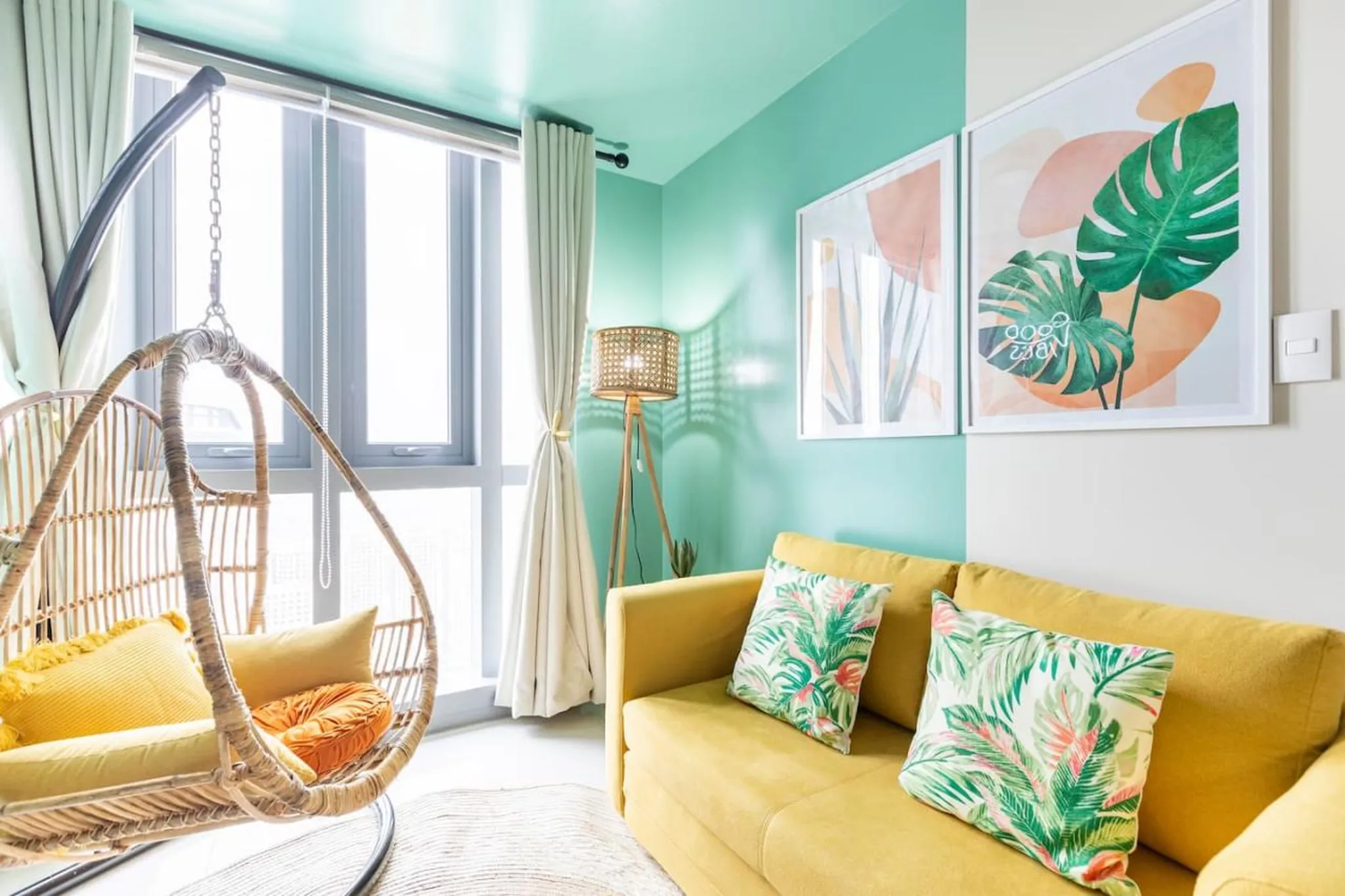 A bright, modern living room with a yellow couch, hanging chair, and tropical-themed art.
