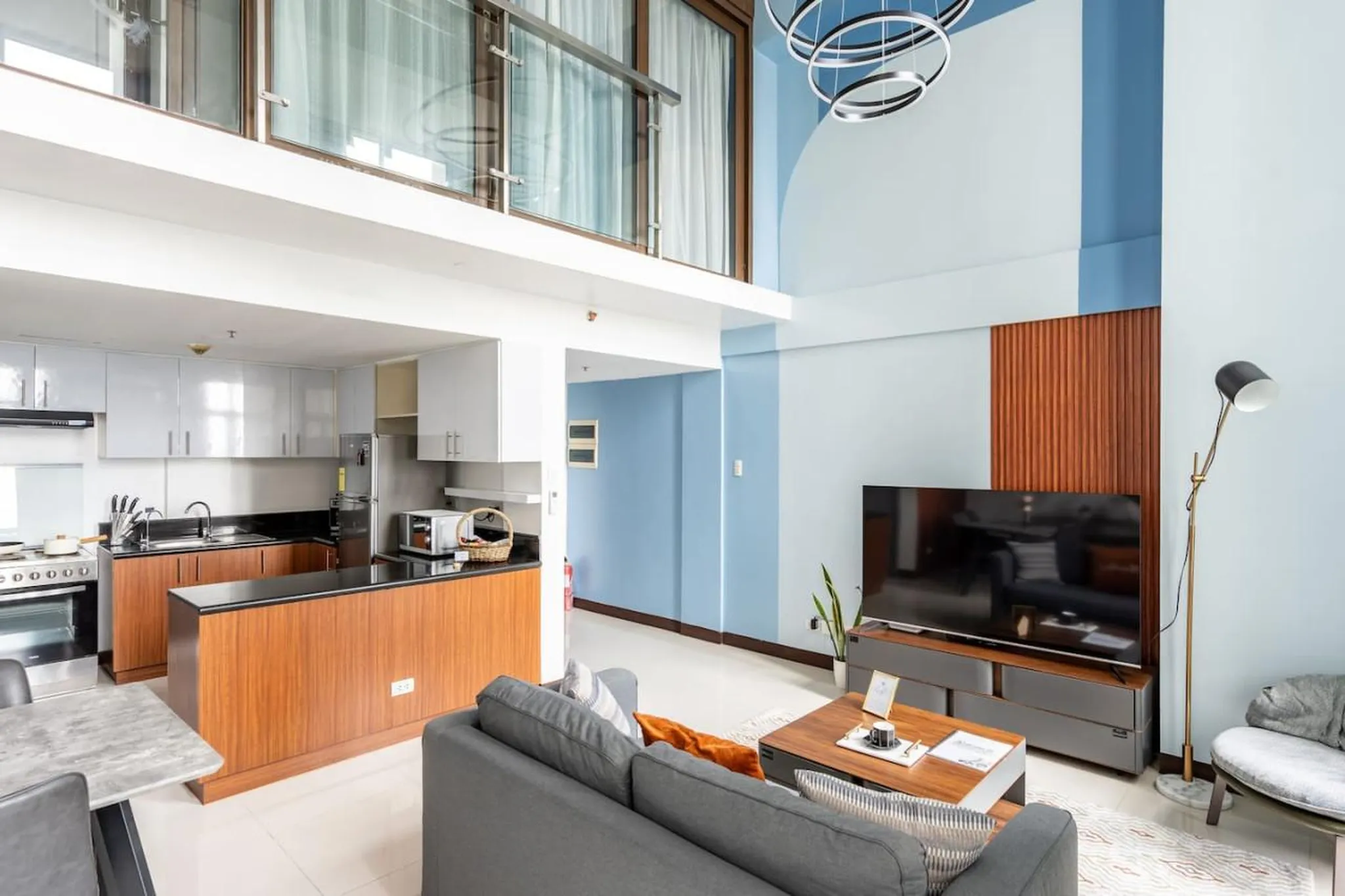 A spacious living area with a modern kitchen, high ceilings, a television, and a grey sofa.