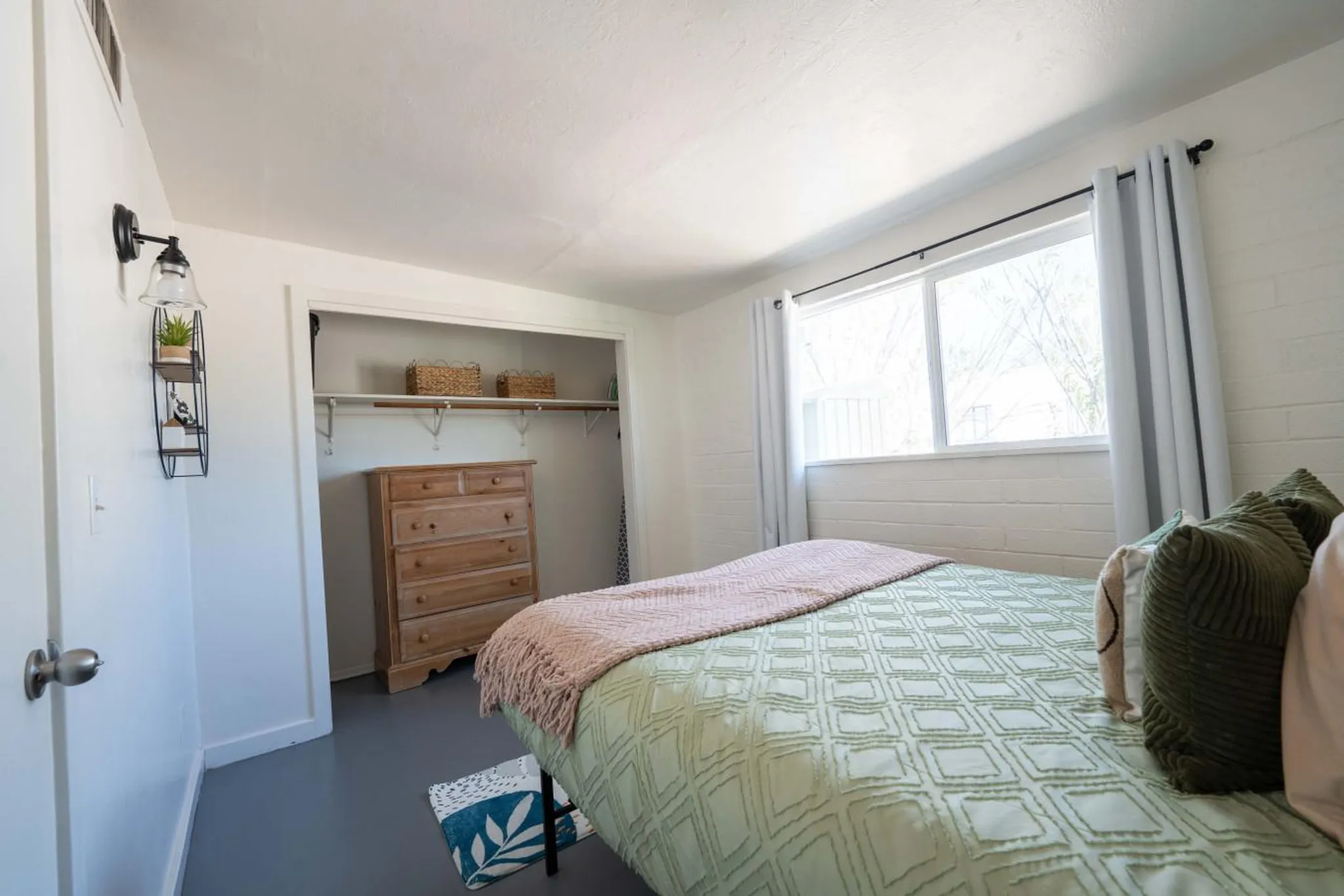 A bedroom with a dresser in a closet, a bed with a pink blanket, and a window with white curtains.
