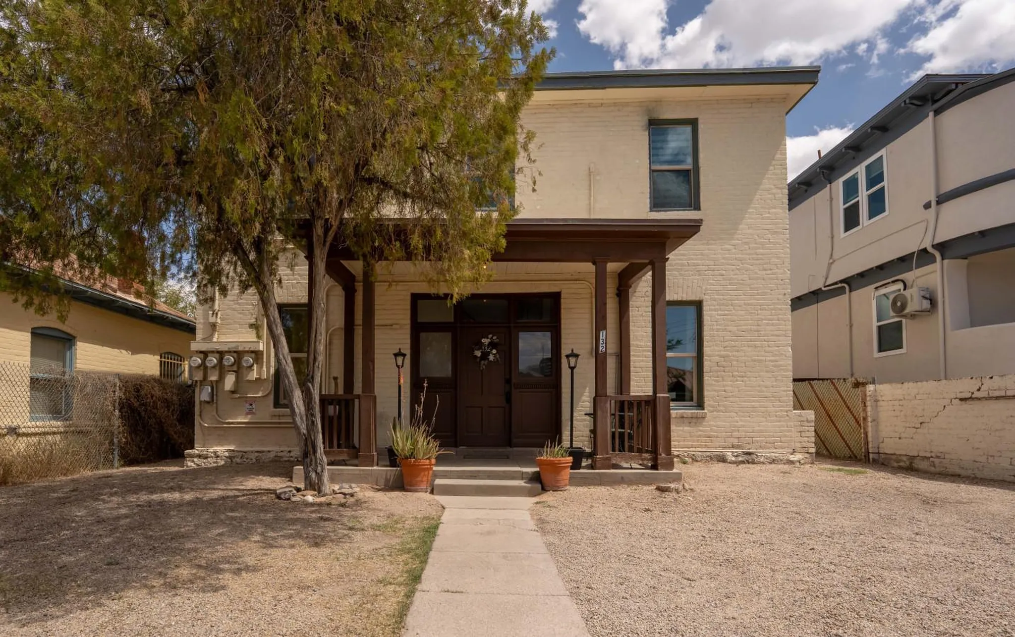 A light yellow brick house with a dark brown porch and door, flanked by potted plants.