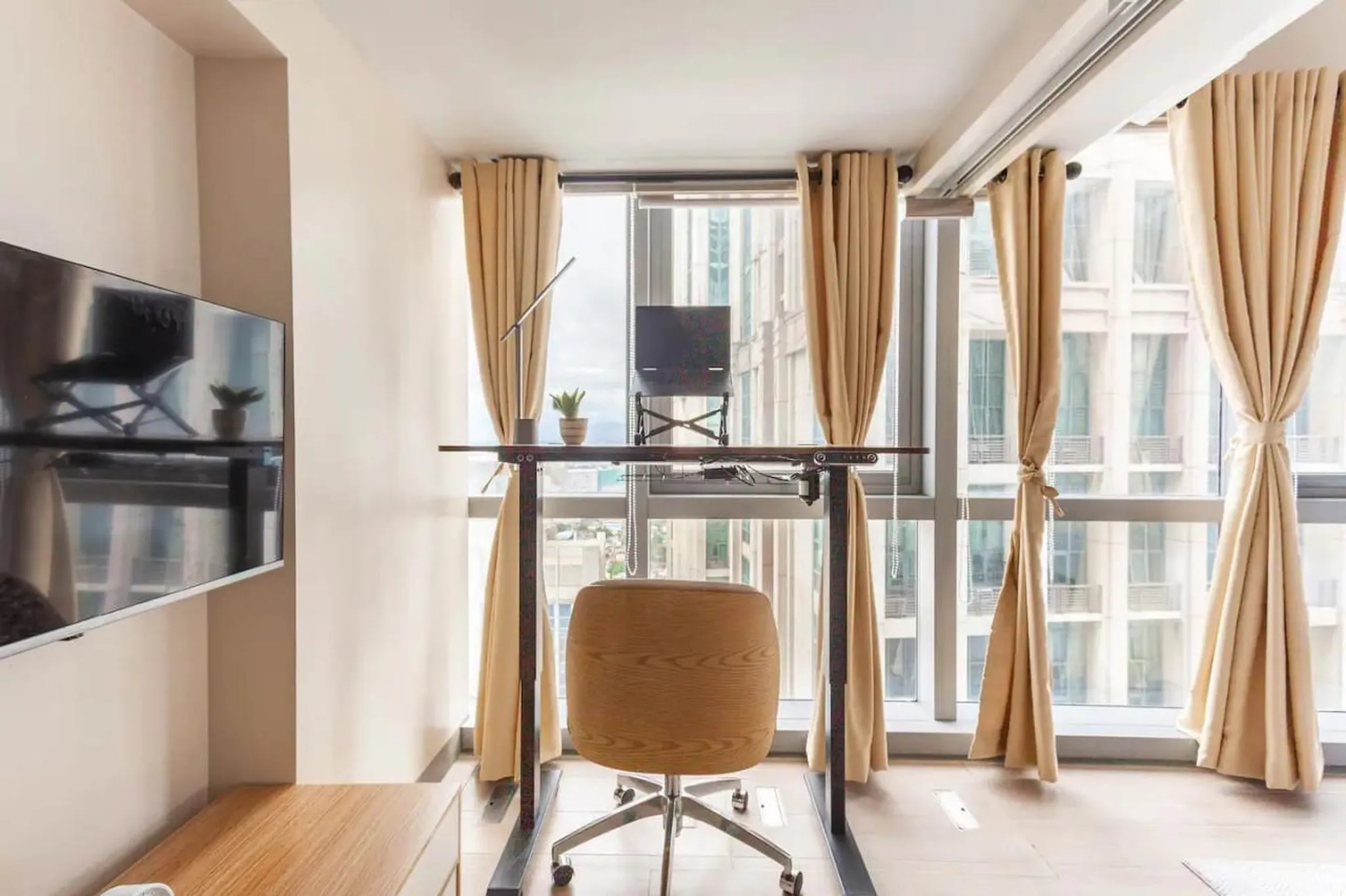 Modern home office with a standing desk, chair, and TV against a wall with large windows.