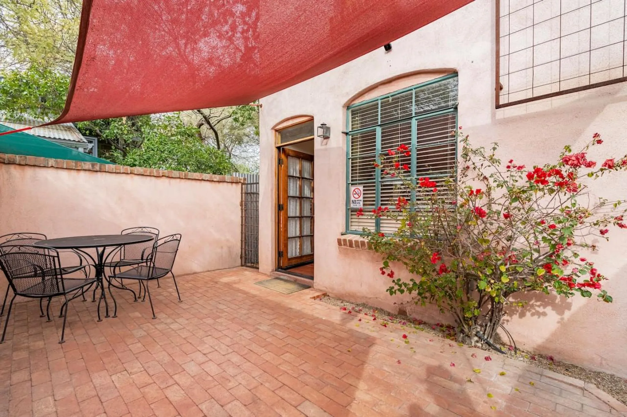 Patio with a table and chairs under a red shade cloth, near a flowering bush.