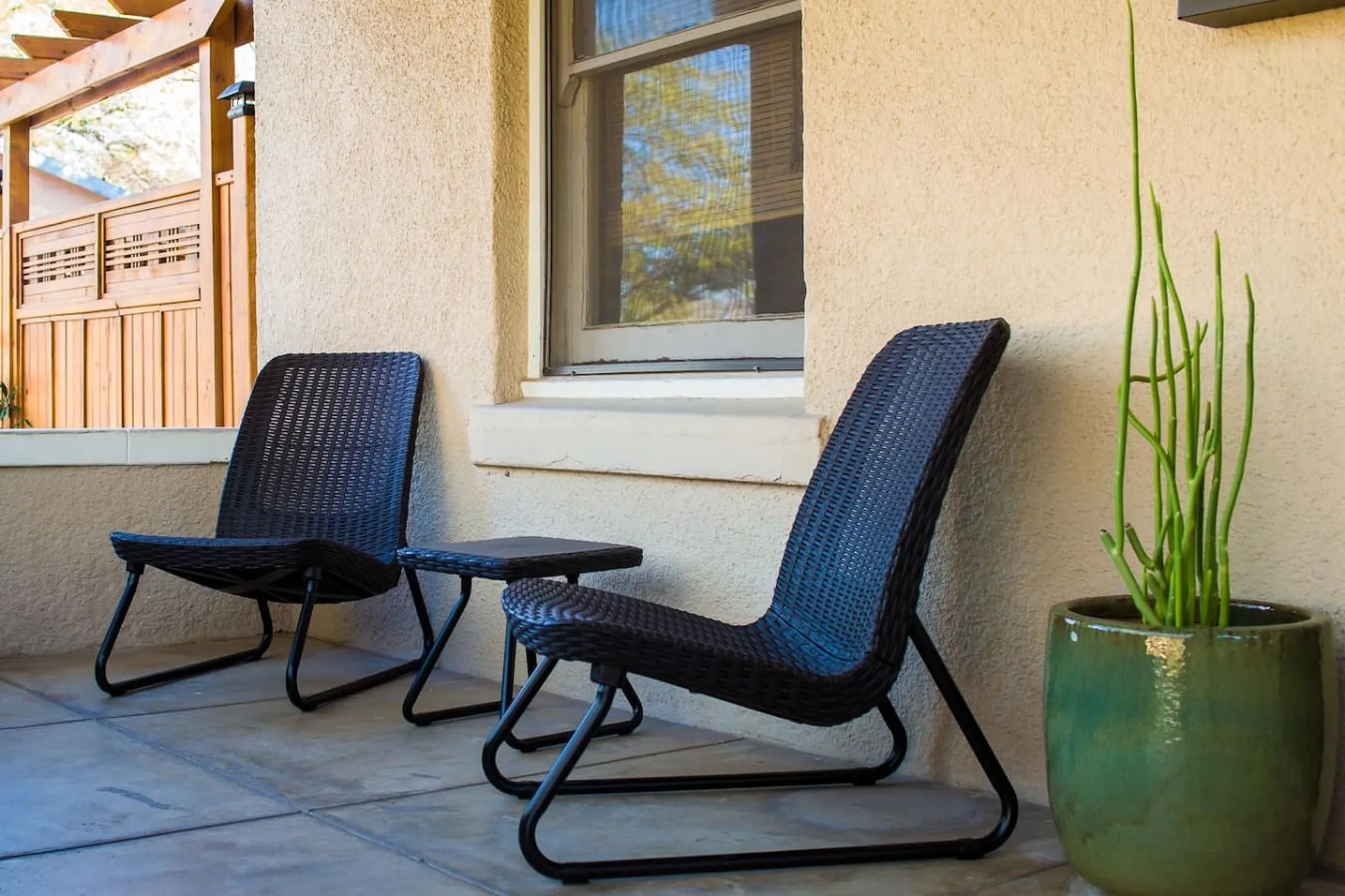 Two dark wicker chairs with a small table on a patio, next to a green pot with a tall green plant.