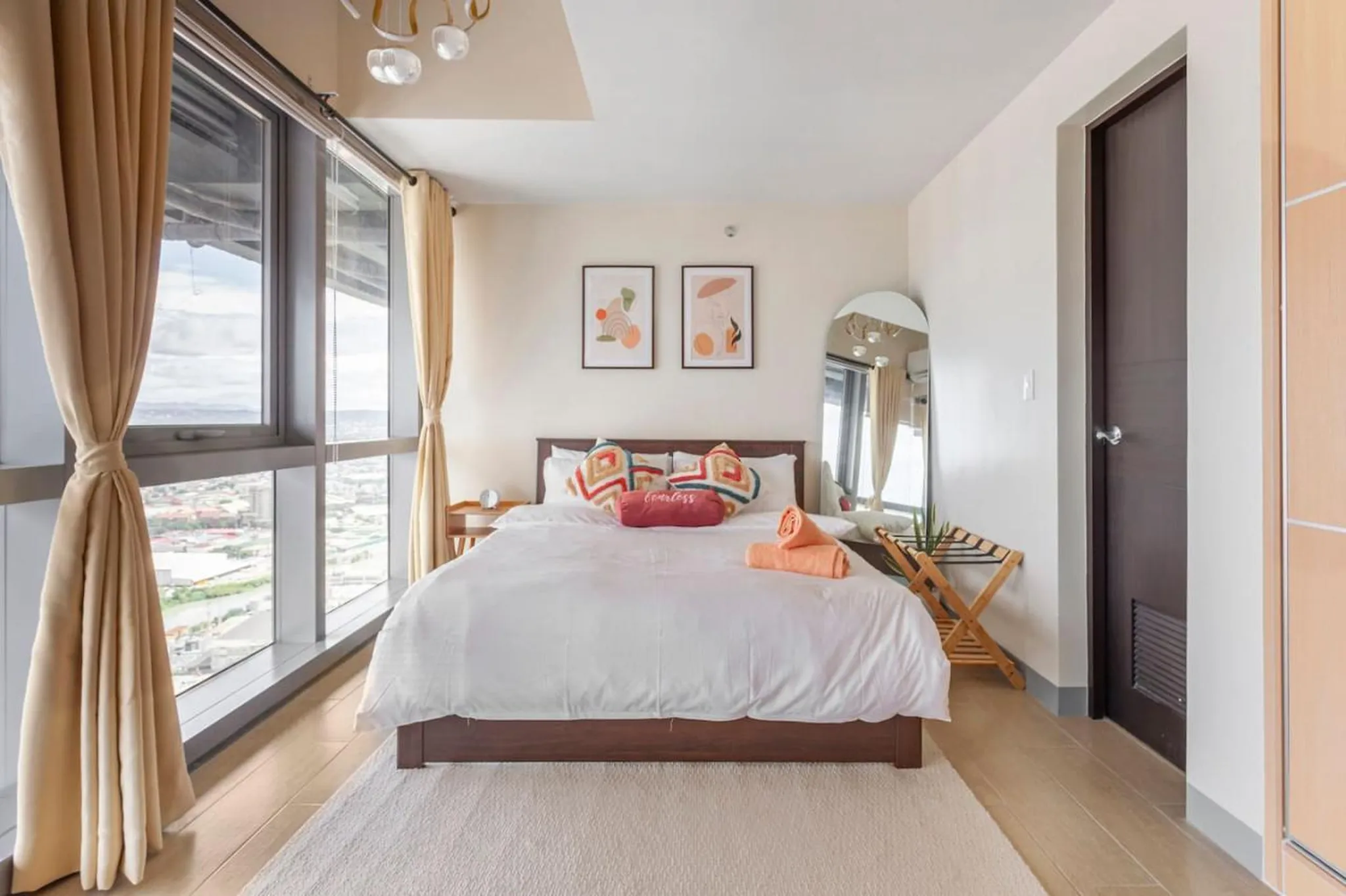 Modern bedroom with large windows and city views, featuring a king-size bed and decorative pillows.