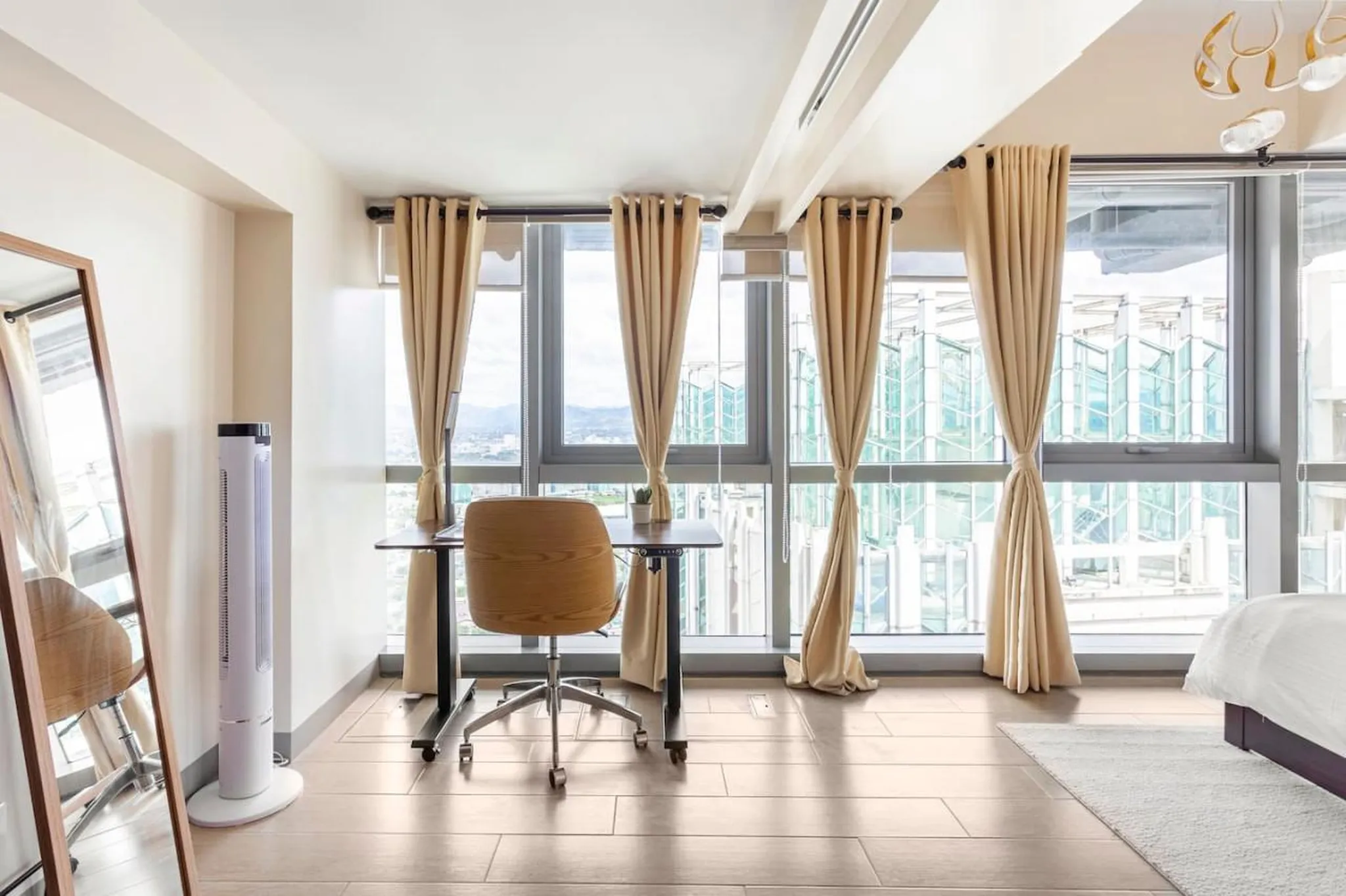 A modern office with a tower fan, desk, chair, and large windows.
