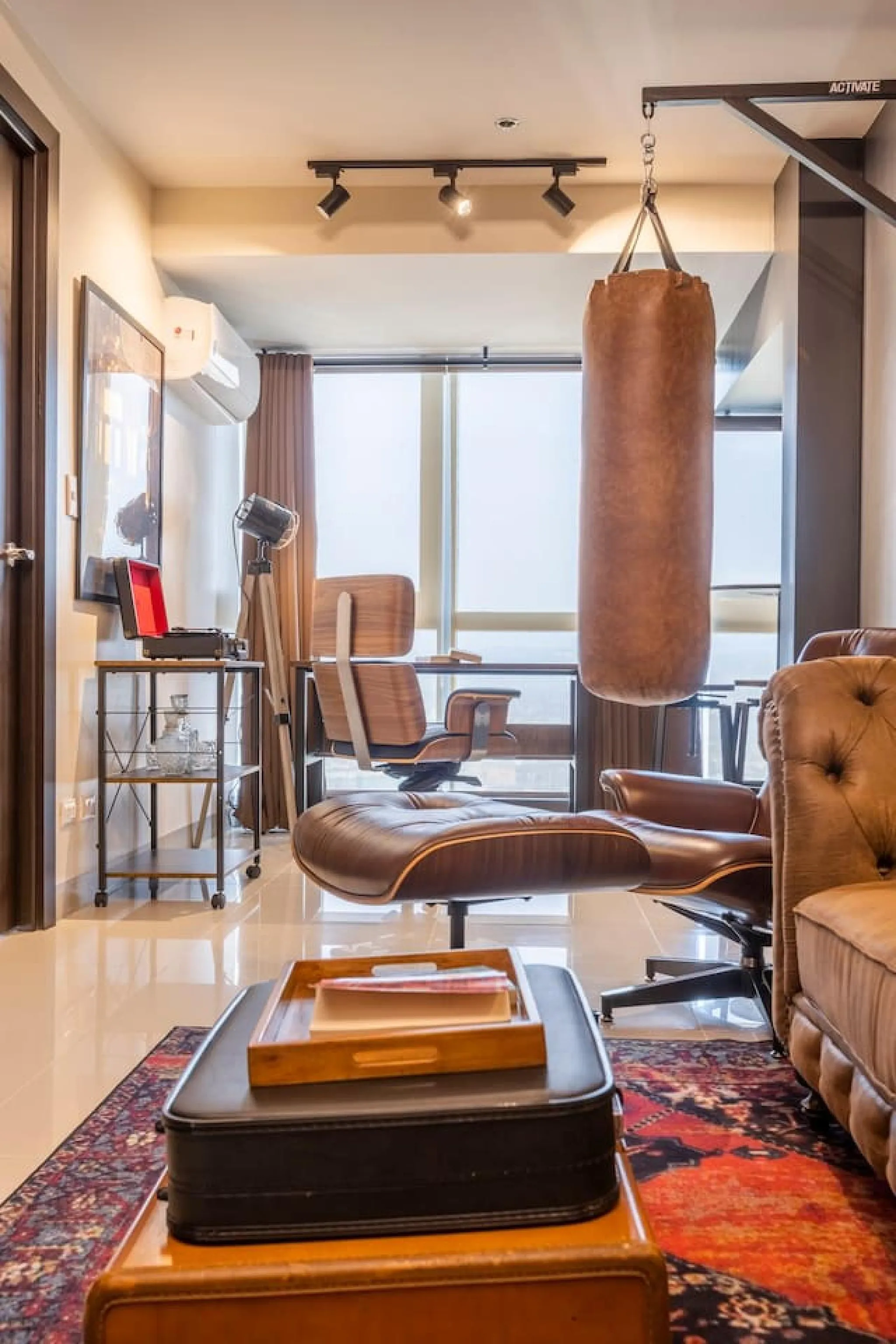 A brown leather punching bag hangs from the ceiling in a room with a desk, chair, and sofa.