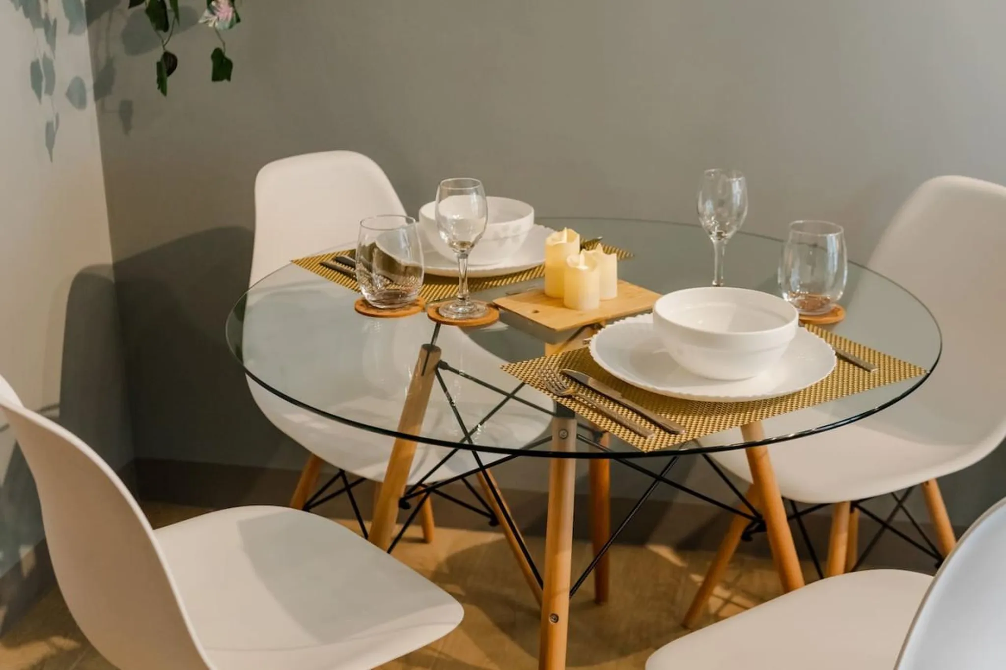 A round glass dining table is set for four with white chairs and candles.