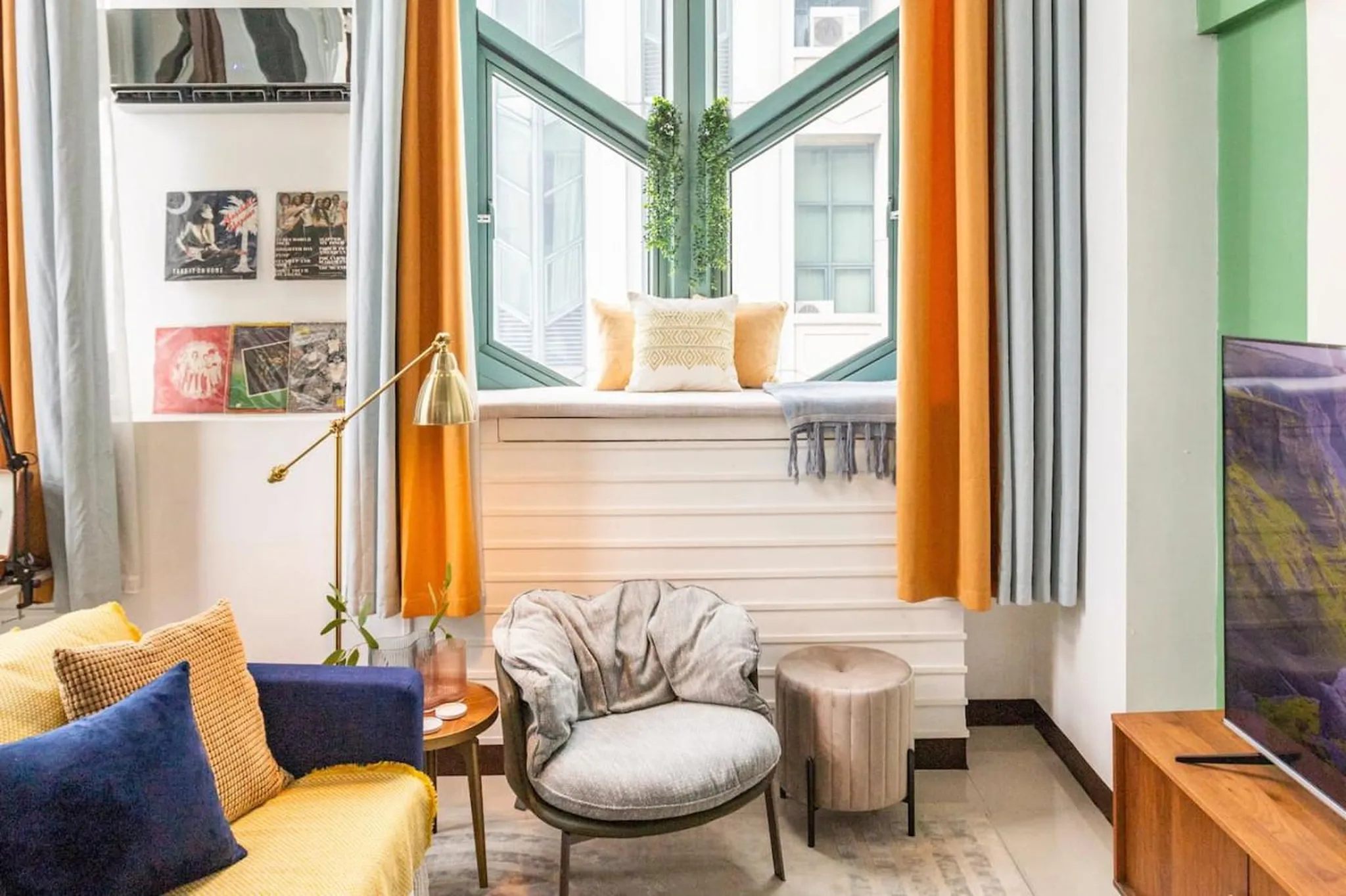 A bright living room with a yellow couch, grey armchair, and a window with orange and blue curtains.