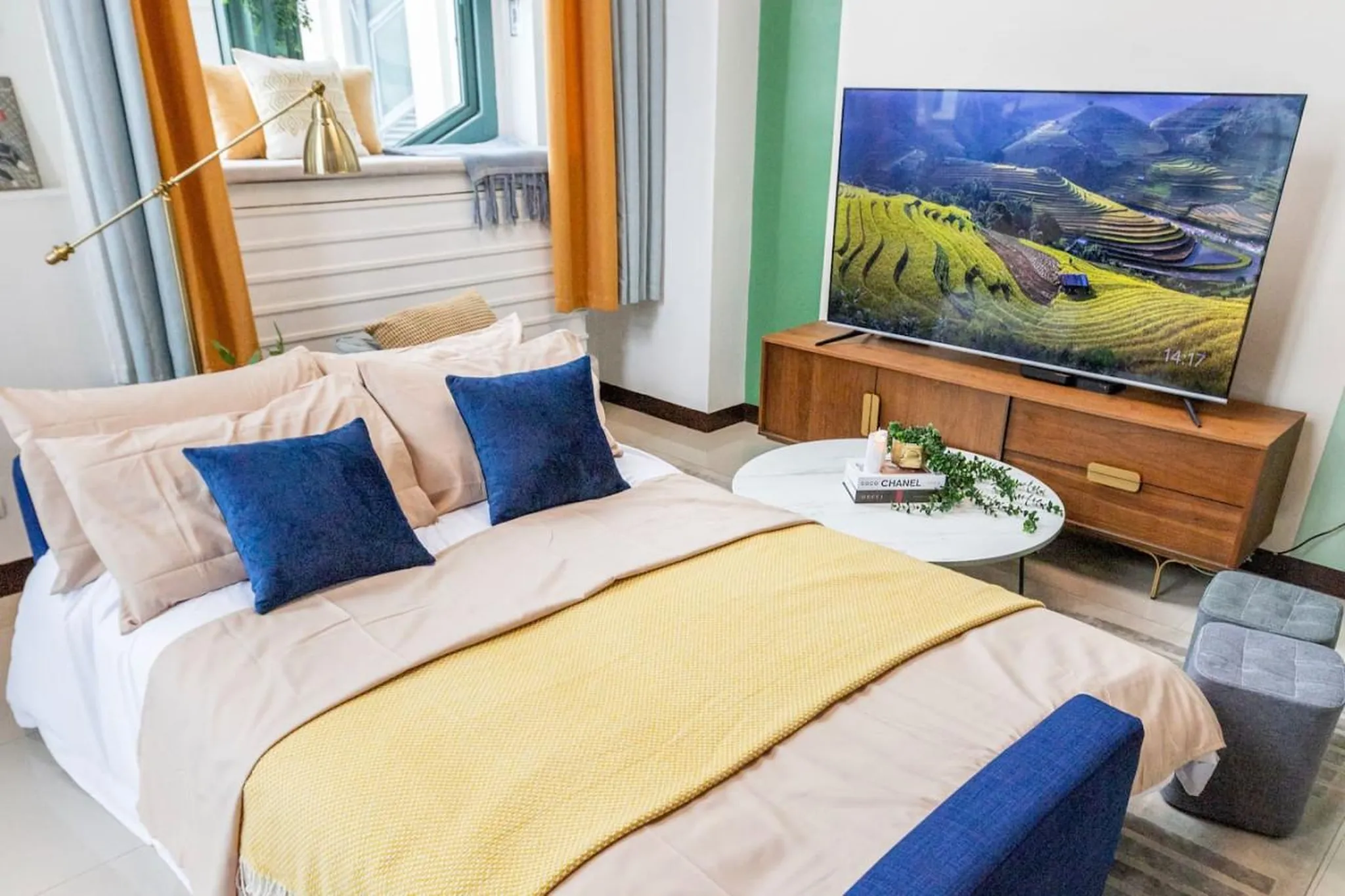 A bed with blue and beige pillows, a yellow blanket, and a TV on a wooden stand.