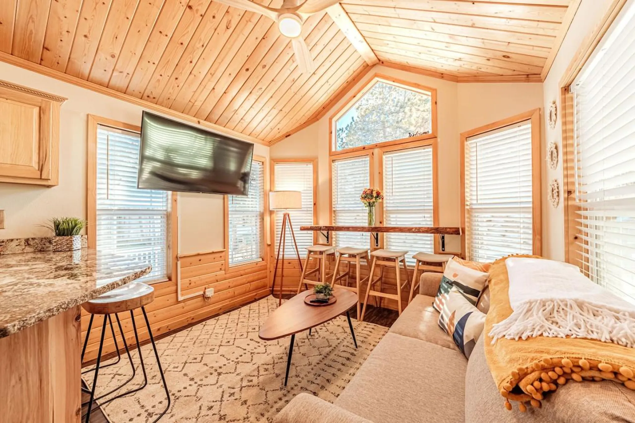 Cozy living area with a TV, sofa, bar, and stools, all in a wood-paneled room with large windows.