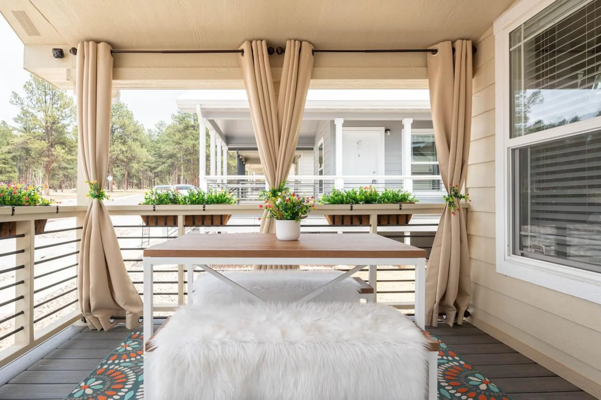 Outdoor patio with beige curtains, a wooden table with a faux fur bench, and flower planters.