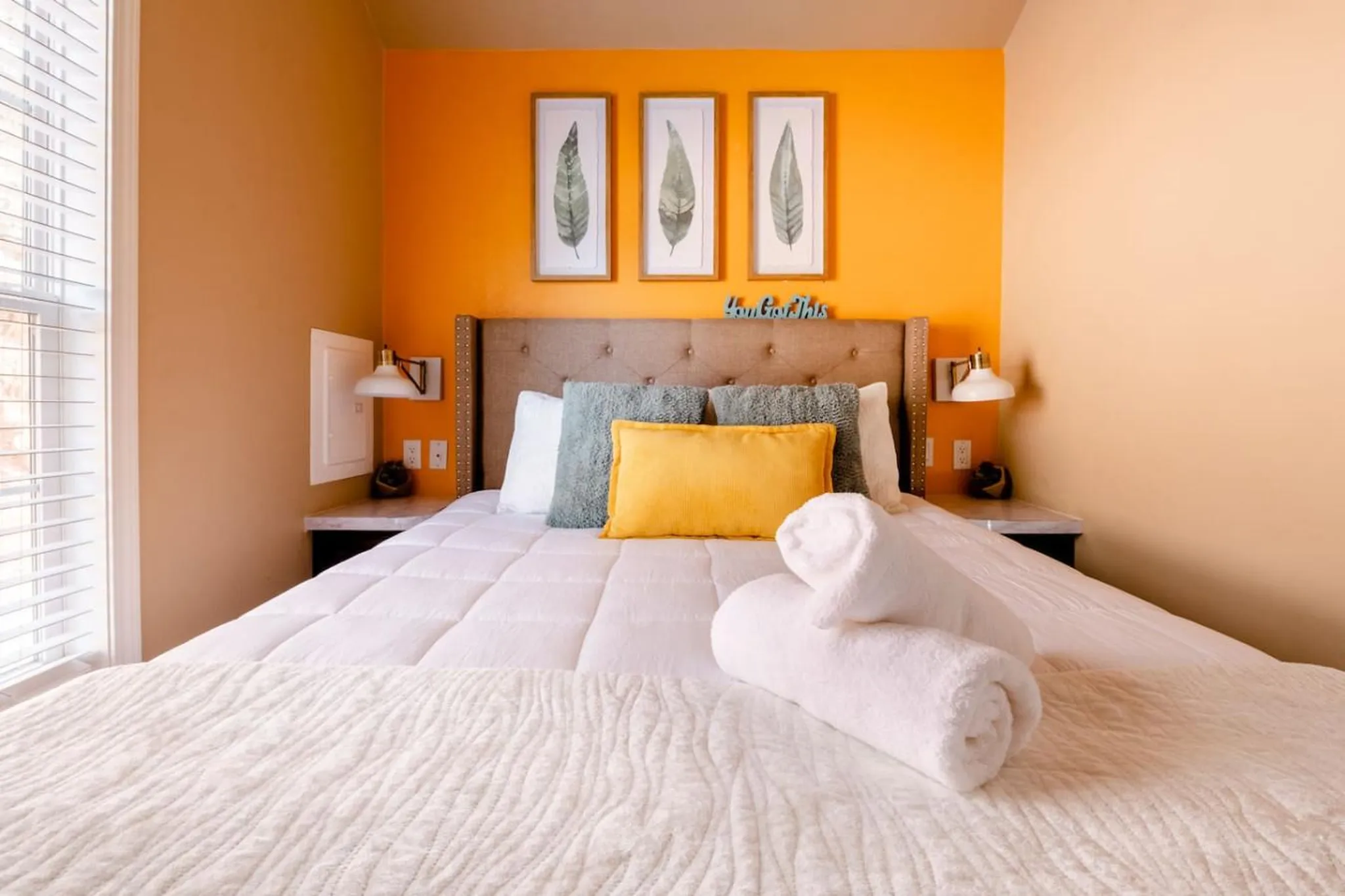 A neatly made bed with white linens and plush pillows, set against an orange wall.