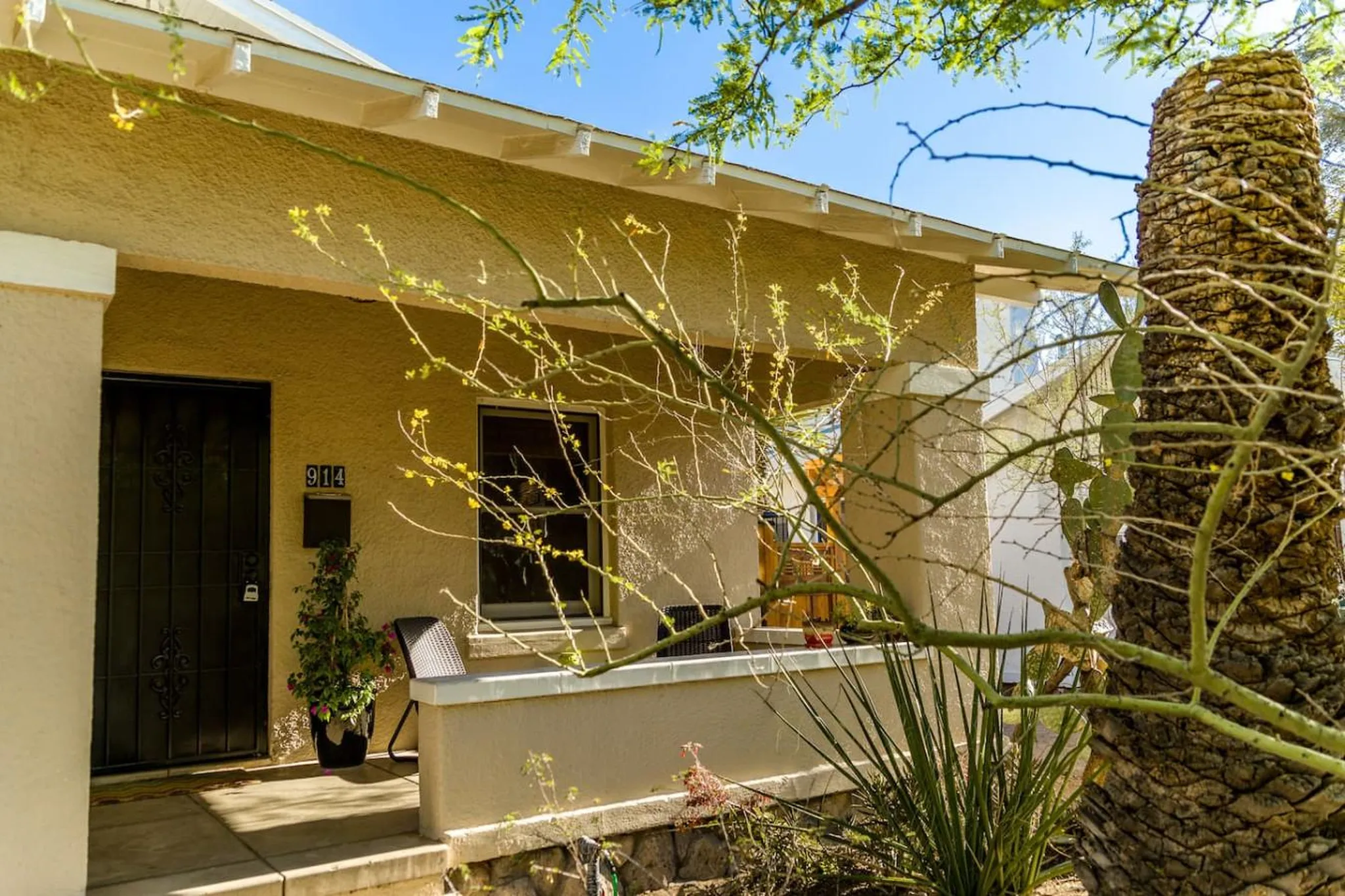 House with stucco exterior, porch, and front door framed by desert plants and trees.