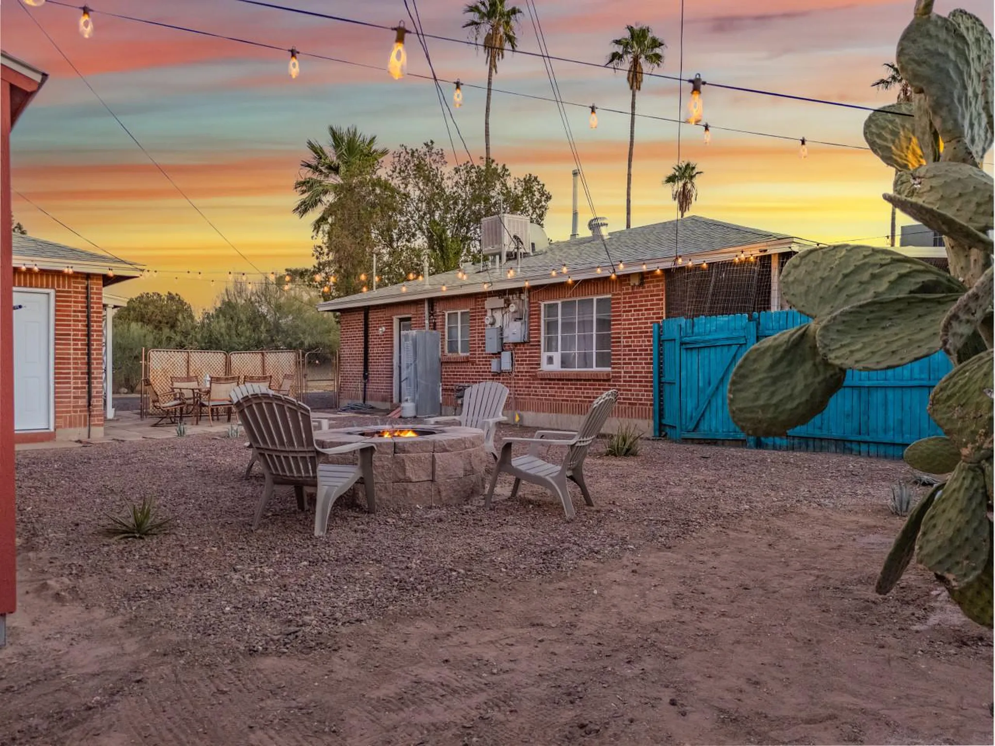 Backyard patio with fire pit, chairs, cactus, and string lights at sunset.