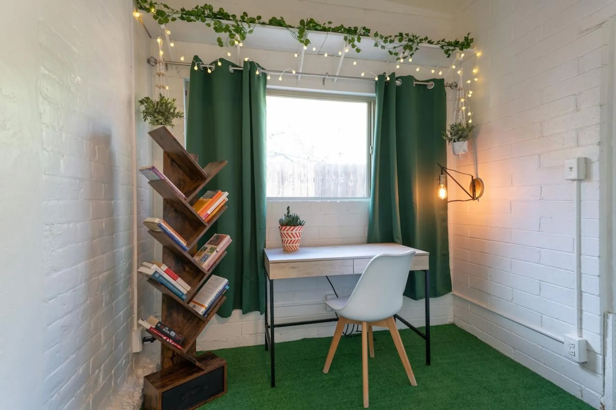 A desk with a white chair is set up next to a window with green curtains in a room with brick walls.