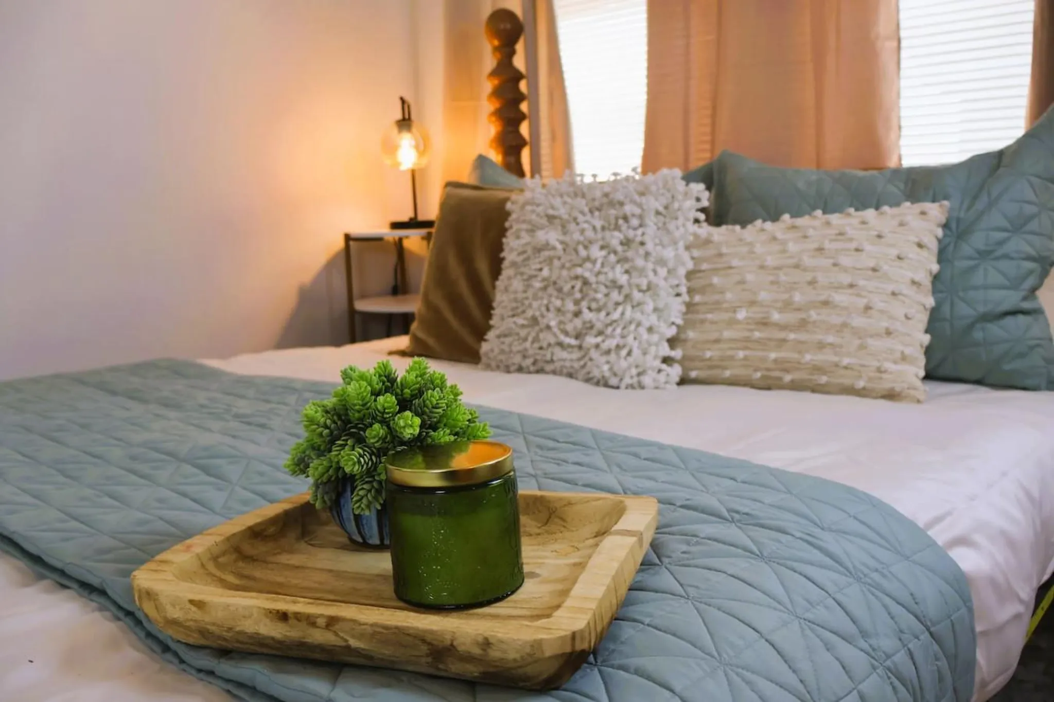 A bed with a light blue quilt, decorative pillows, a plant, and a candle on a wooden tray.