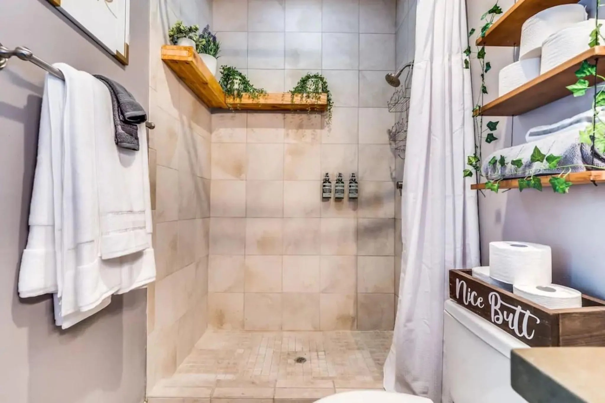 A bathroom with a tiled shower, towels, plants, and toilet paper.
