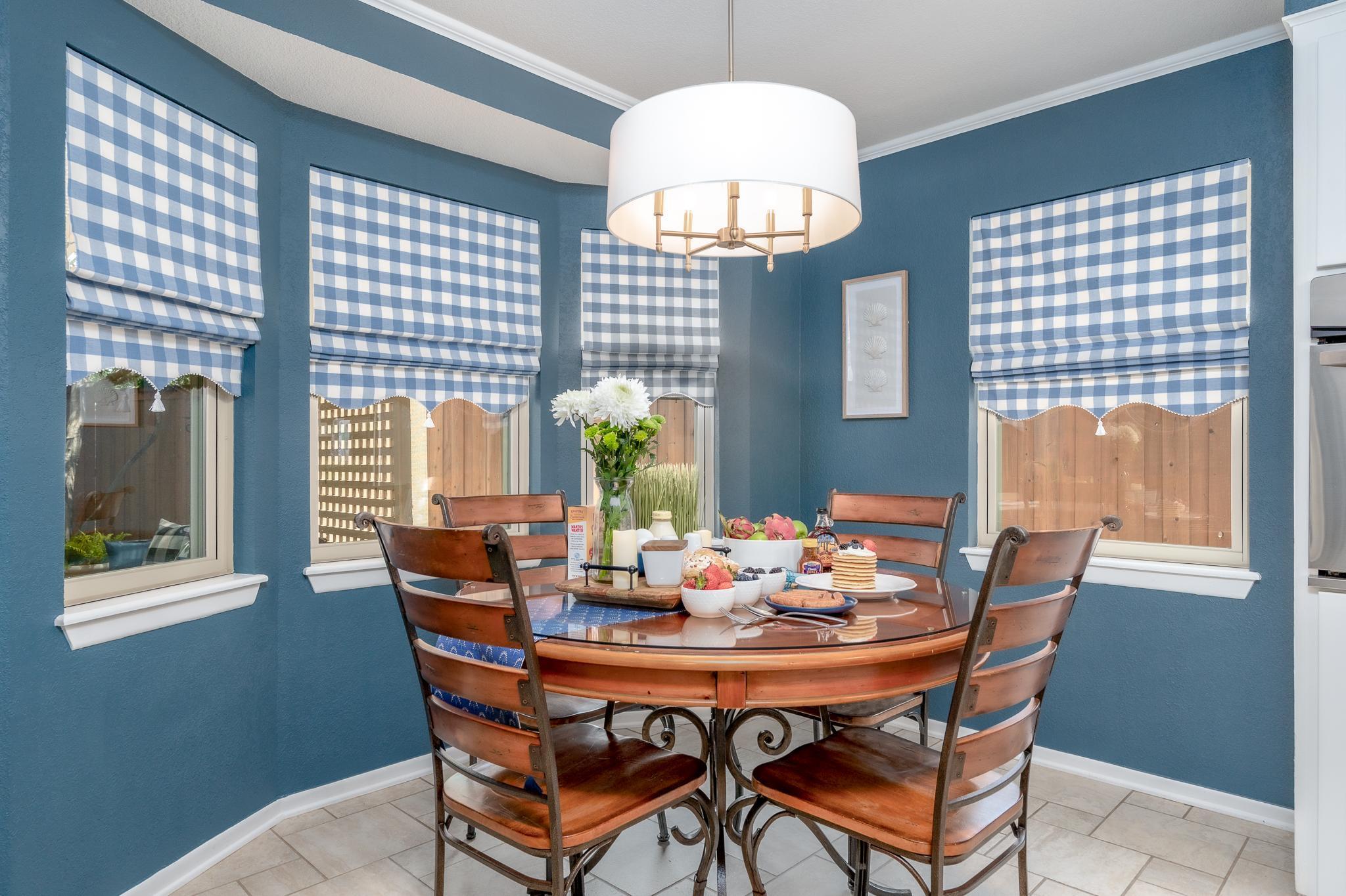 A round dining table is set for breakfast in a room with blue walls and gingham curtains.