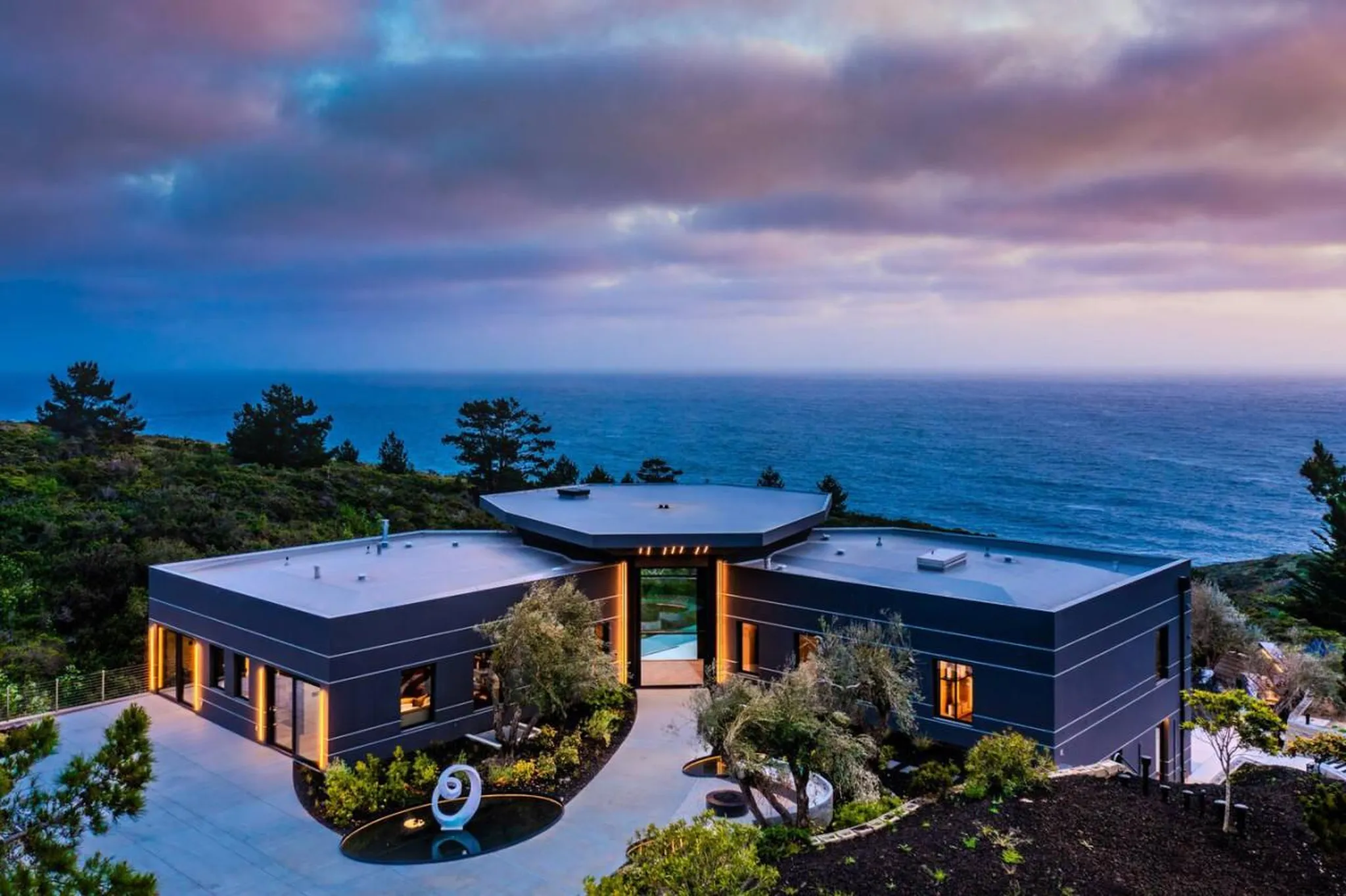 Modern dark gray house with a multi-faceted roof sits on a cliff overlooking the ocean at dusk.