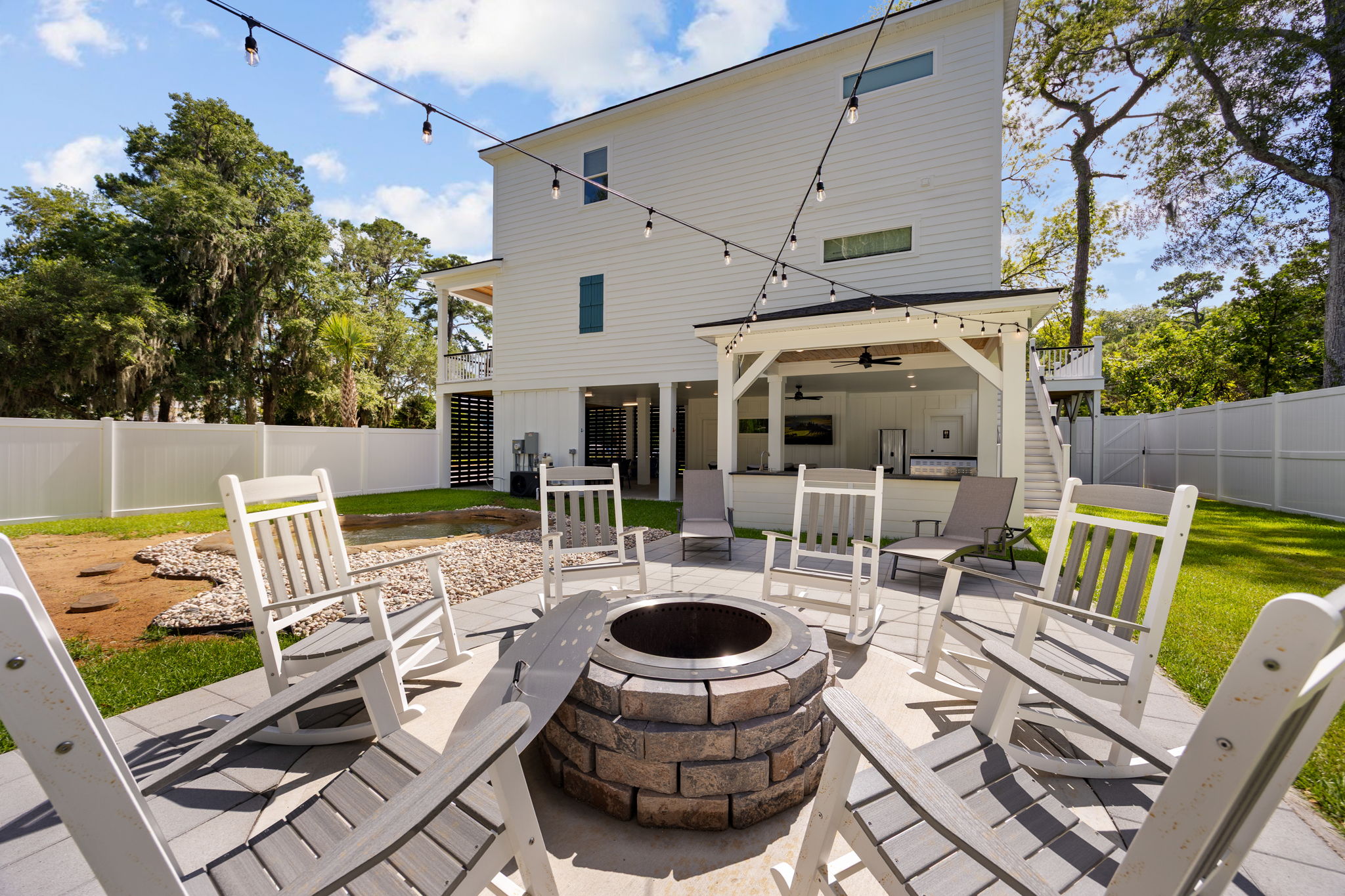Backyard patio with circular stone fire pit surrounded by white rocking chairs, string lights, and a covered outdoor kitchen area